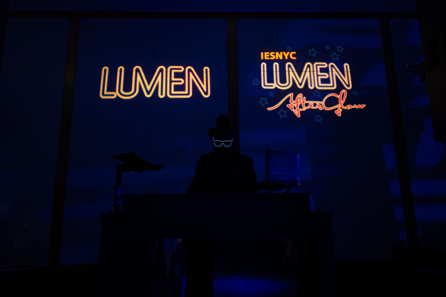A silhouetted DJ performs beneath glowing LUMEN IESNYC neon signs in a darkened venue.