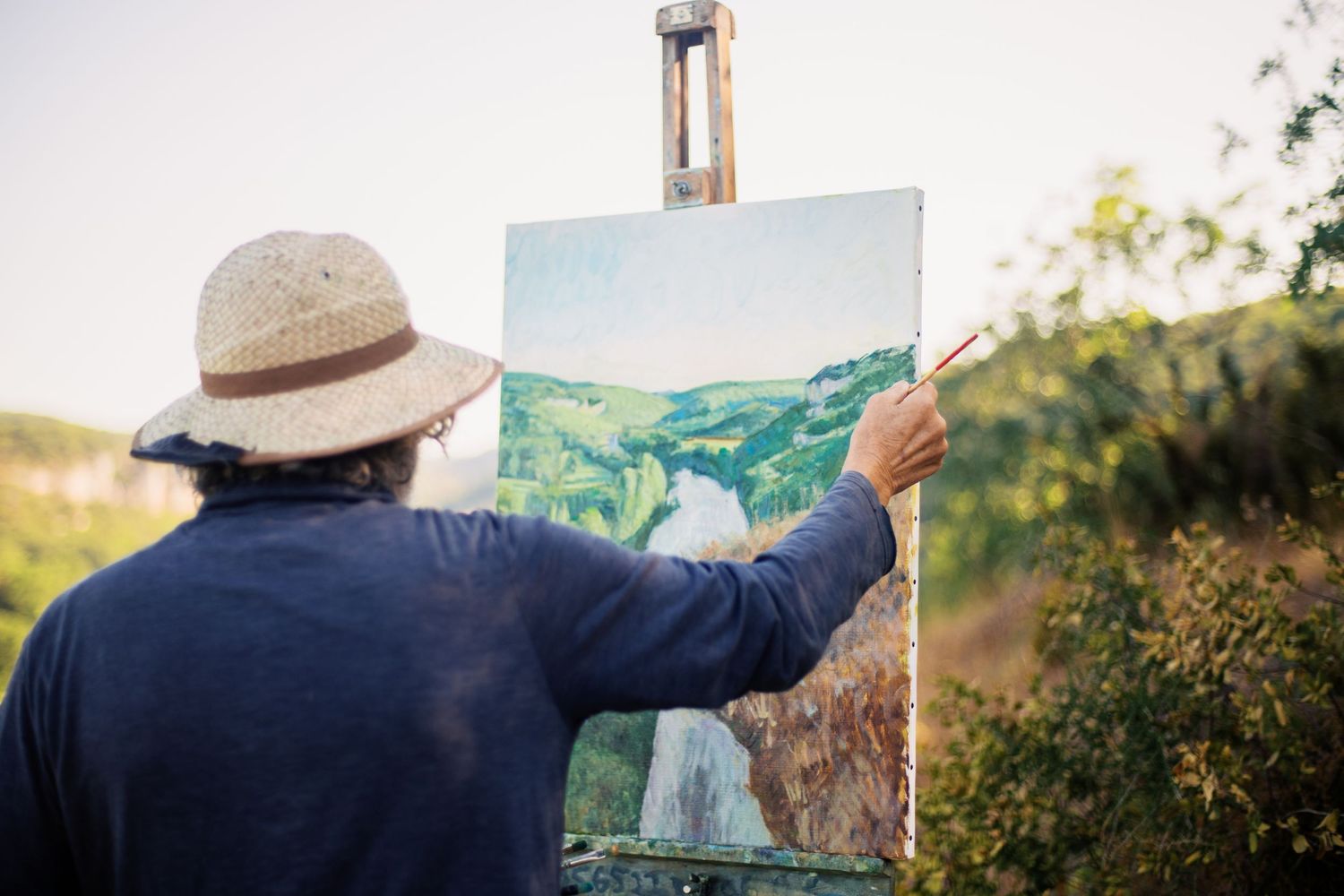 Artist painting a landscape on canvas outdoors on an easel, wearing a straw hat in a sunny garden setting.