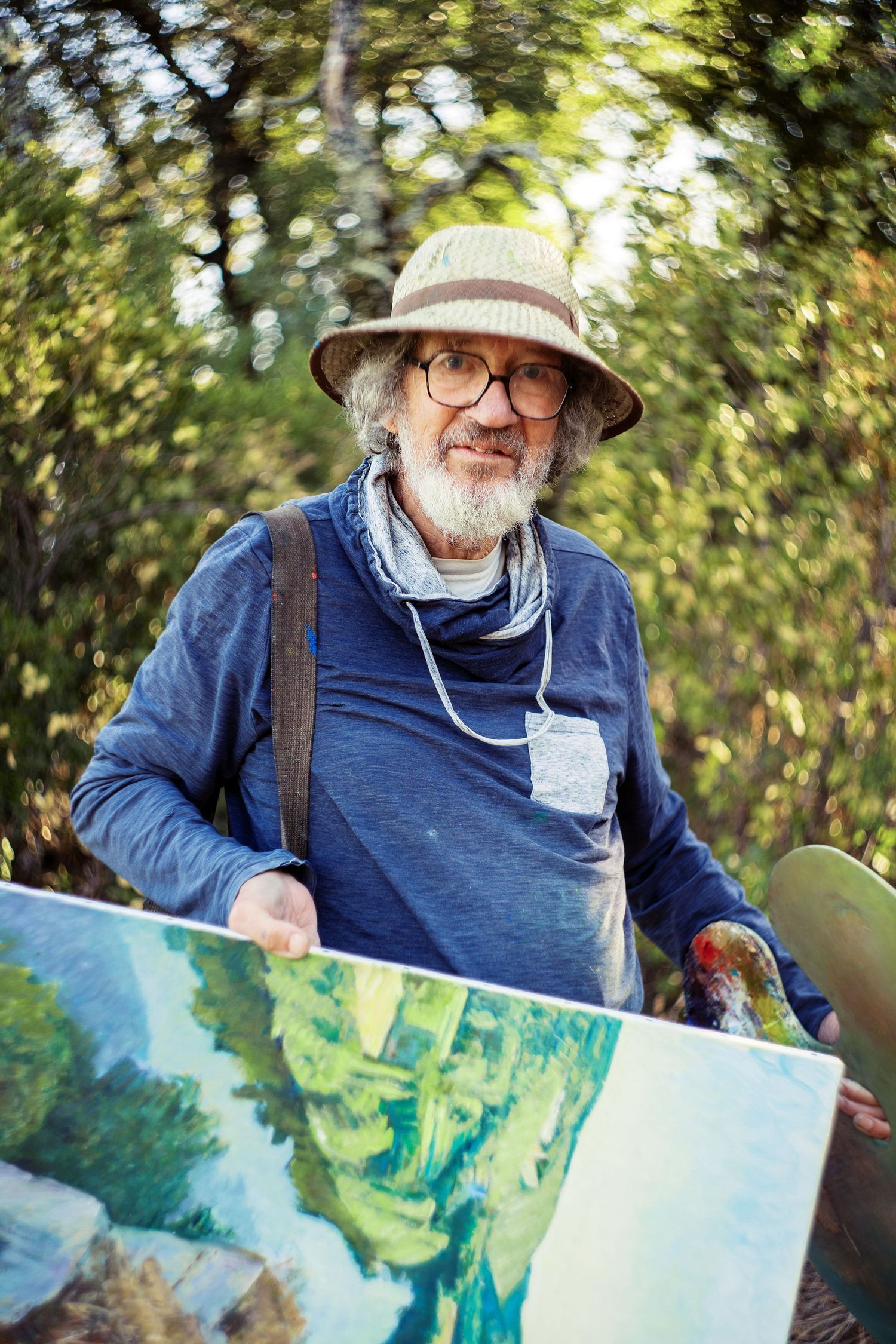 An elderly male artist with glasses and a hat holds a colorful landscape painting outdoors among green trees.