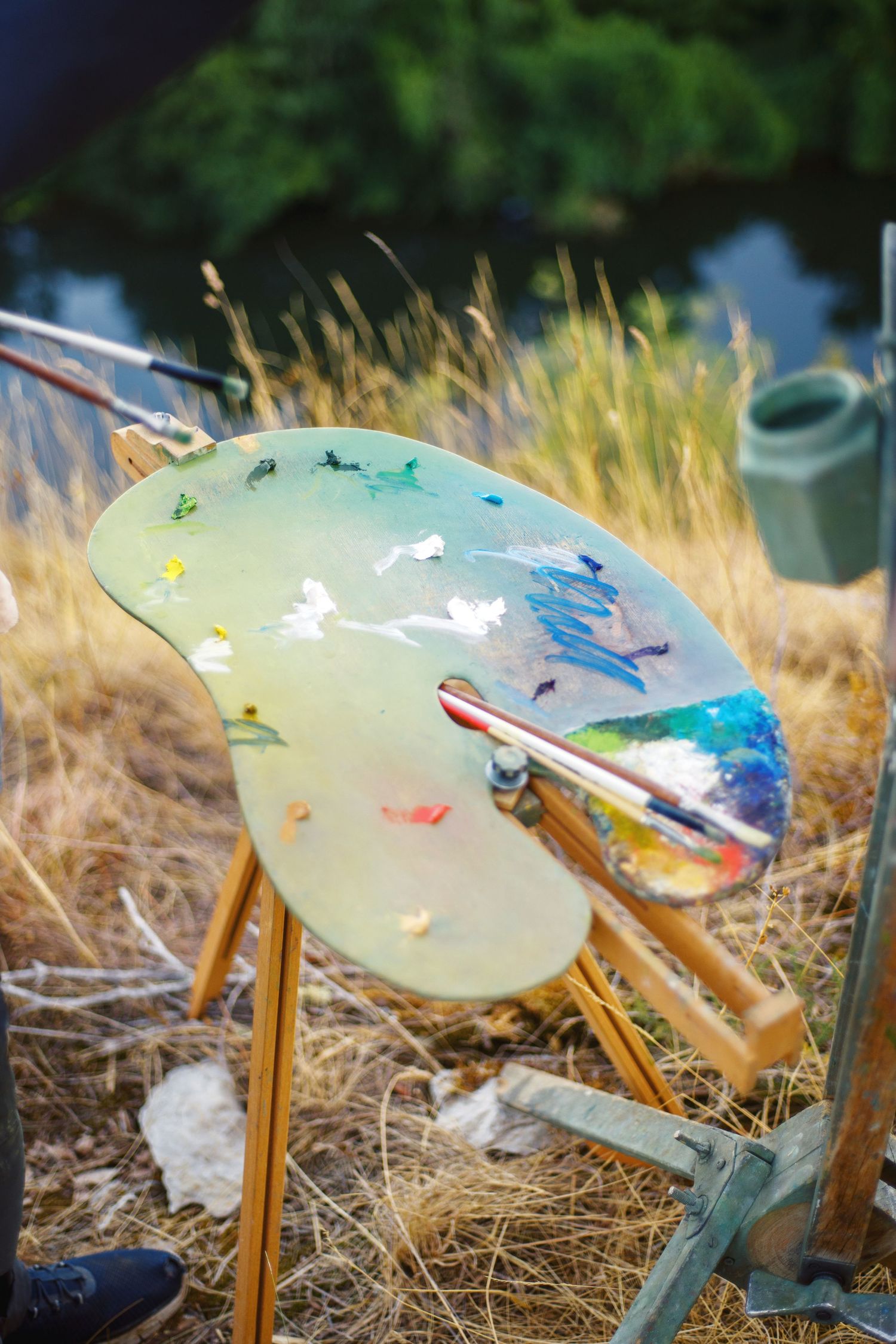 Artist's paint palette with colorful oil paints and brushes resting on an easel outdoors in a grassy field.