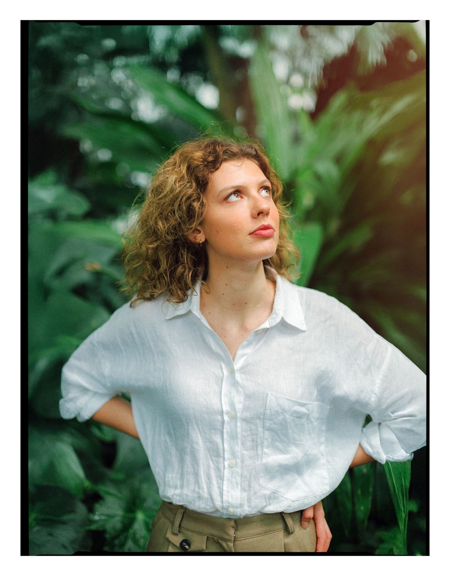 Young woman with curly hair in white linen shirt poses confidently among lush tropical green foliage.