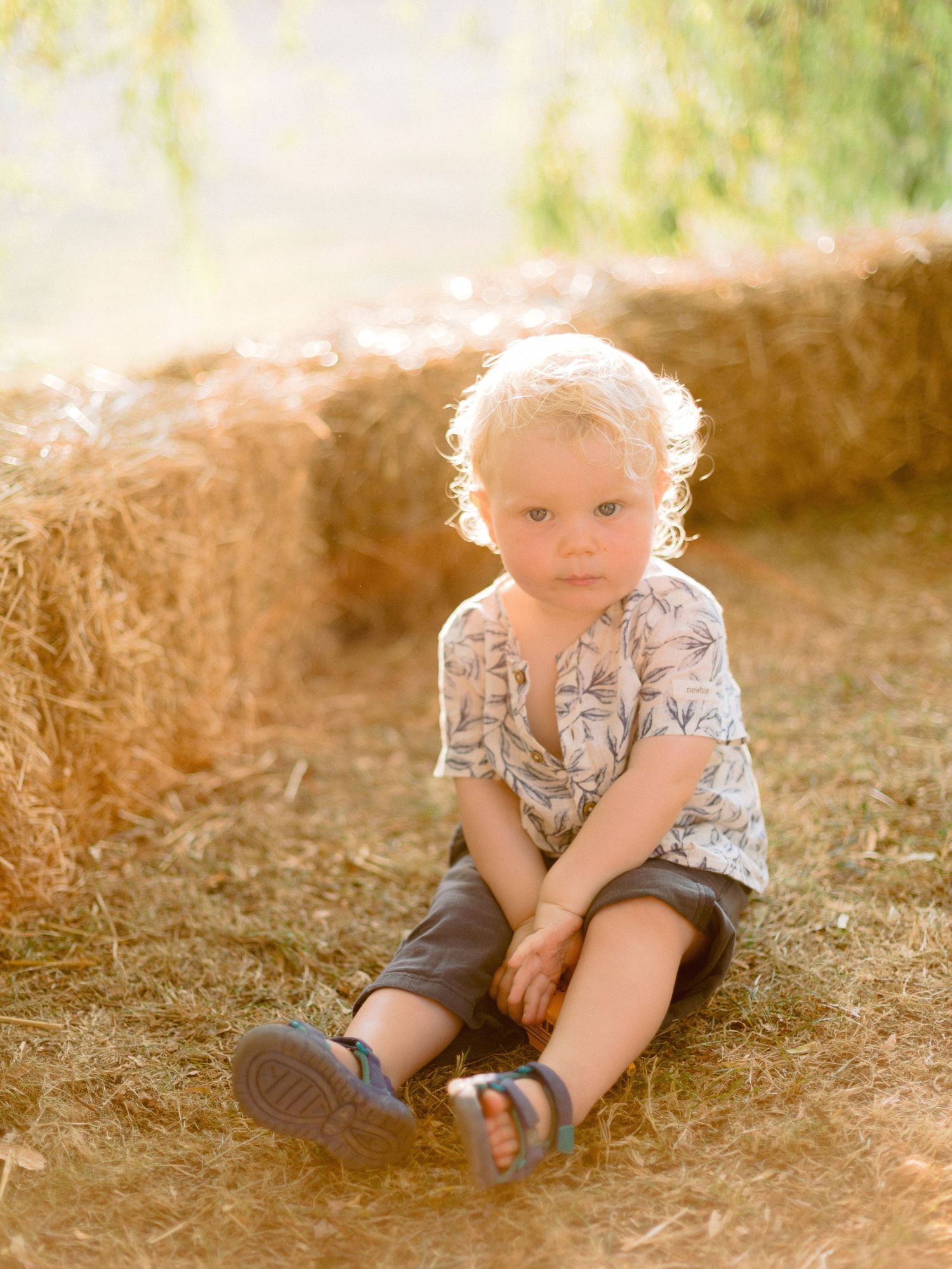 Small child sitting on ground wearing floral shirt and shorts in warm natural sunlight with hay bale in background.