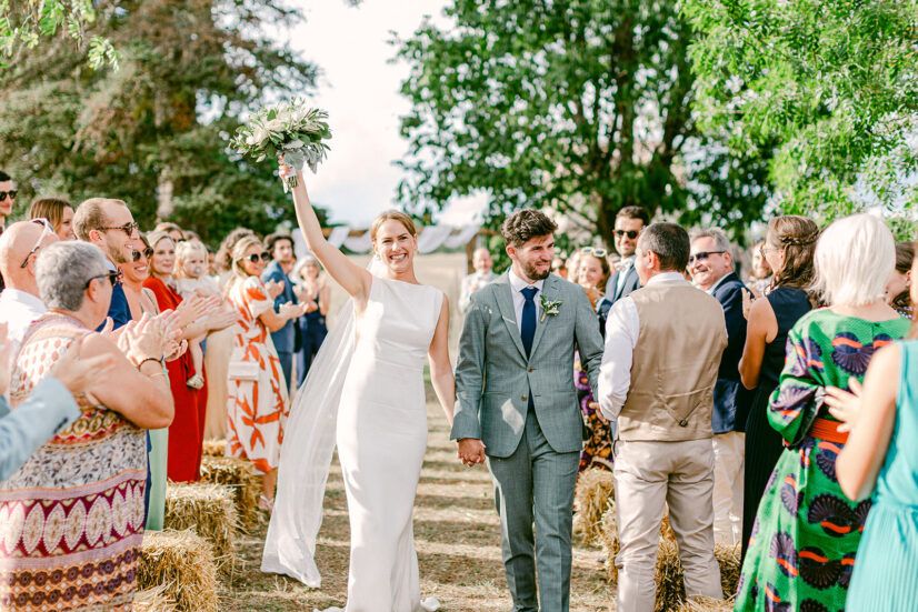 Bride and groom walk joyfully down the aisle surrounded by cheering wedding guests outdoors.
