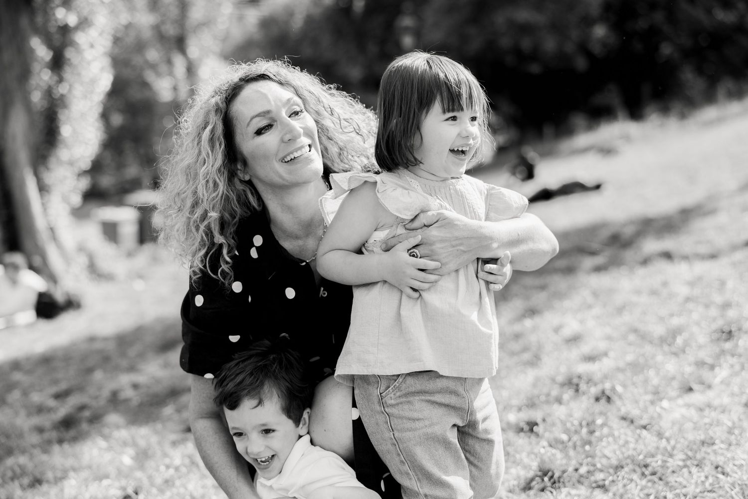 Black and white photo of a smiling woman with curly hair hugging two young children outdoors in a park.