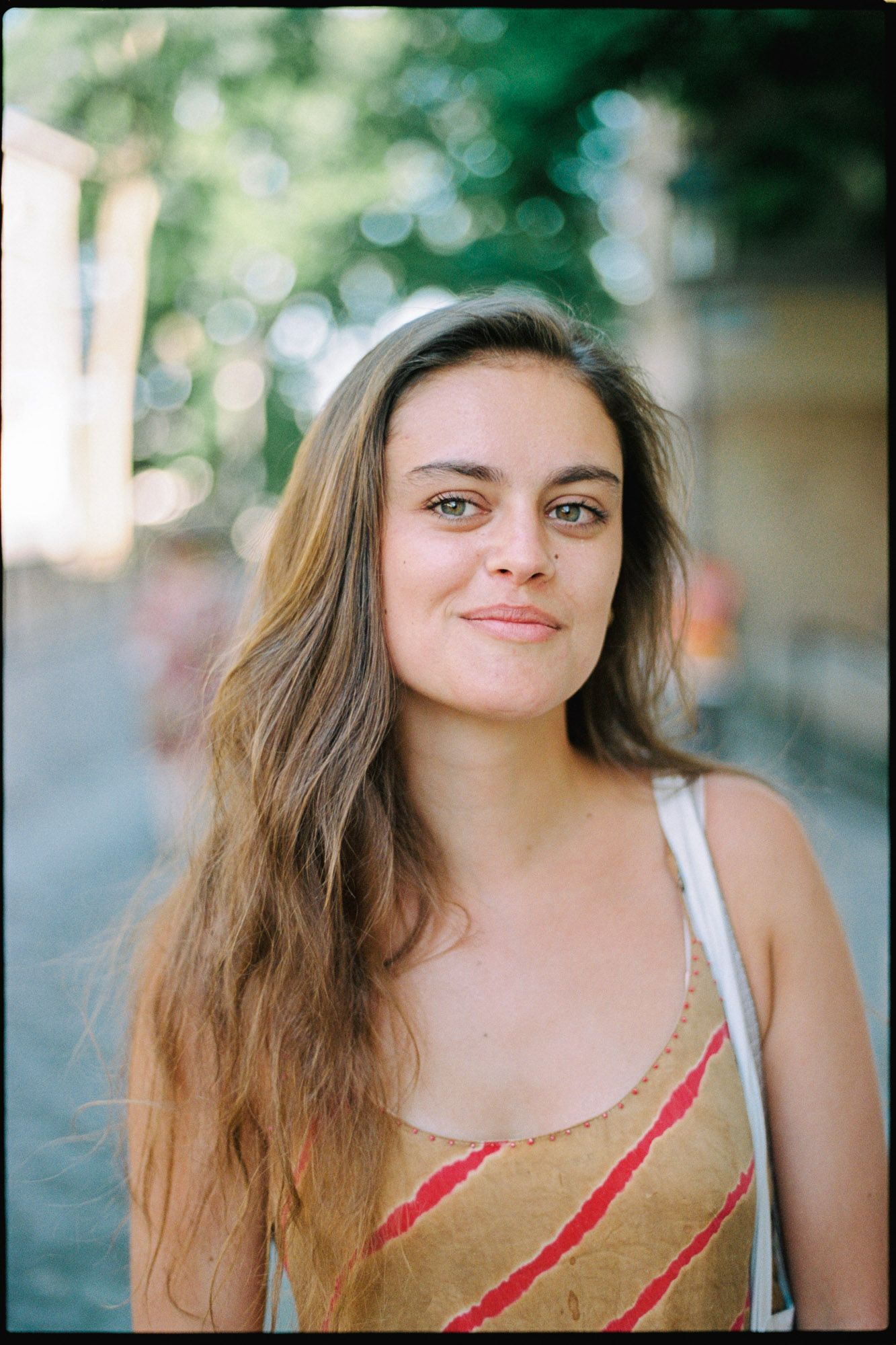 Young woman with long brown hair and freckles smiles softly outdoors, wearing a red and gold sundress.