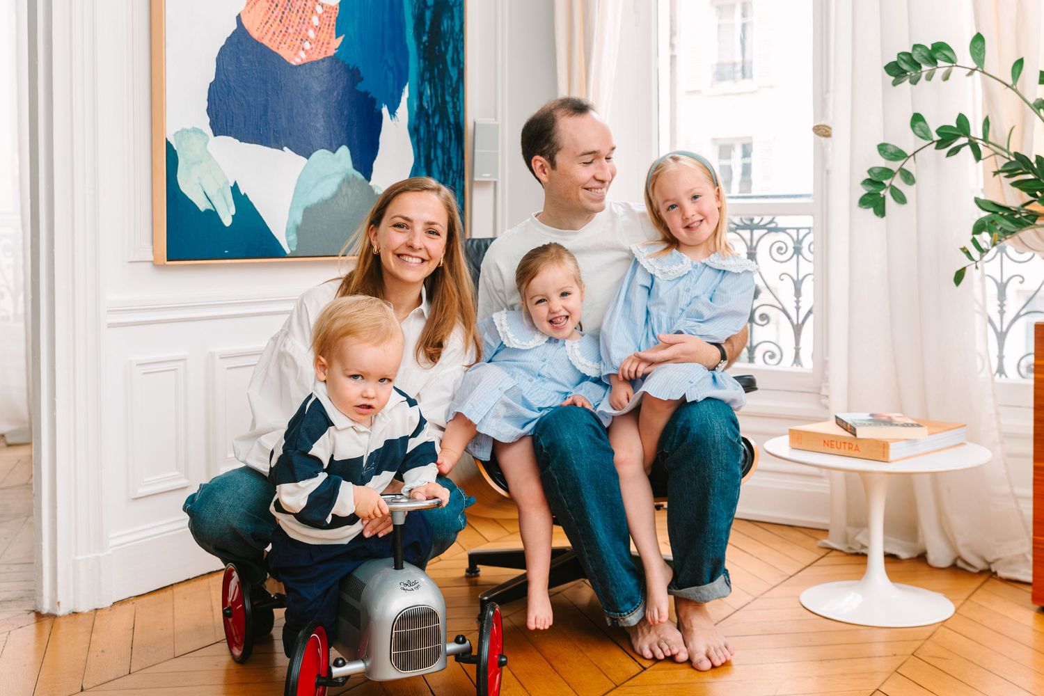 Happy family of five posing indoors, parents with three young children, one riding a toy car in a bright living room.