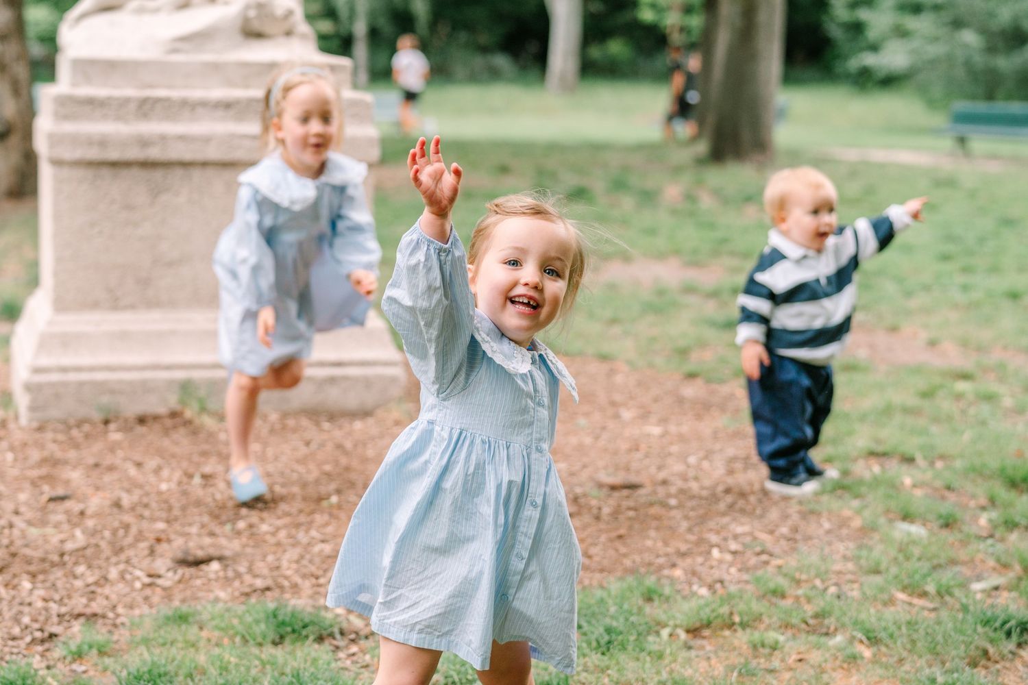 Three young children in matching blue outfits playing and running outdoors in a park near a stone monument.