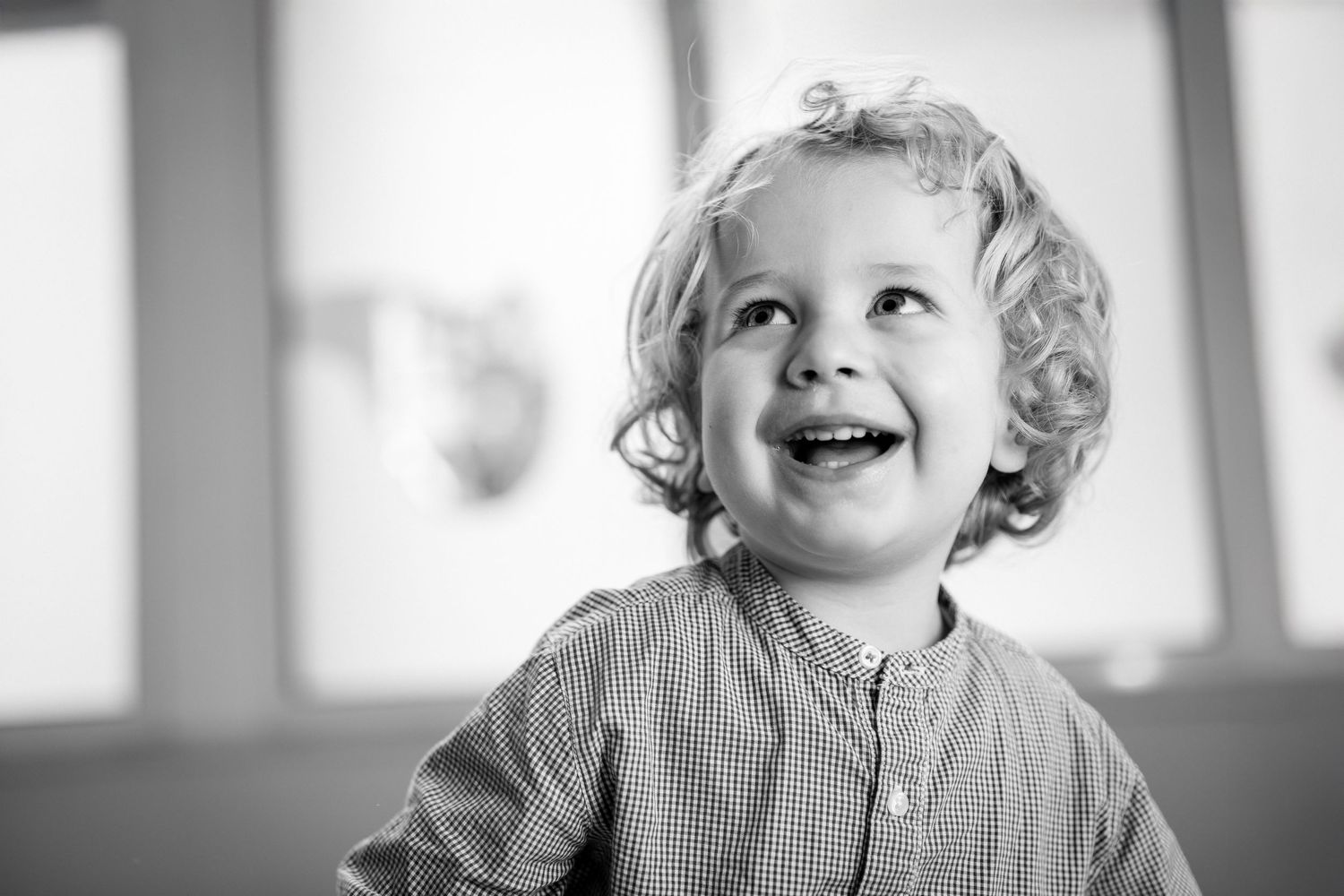 A joyful toddler with curly blonde hair laughs and looks up in a black and white portrait photo.
