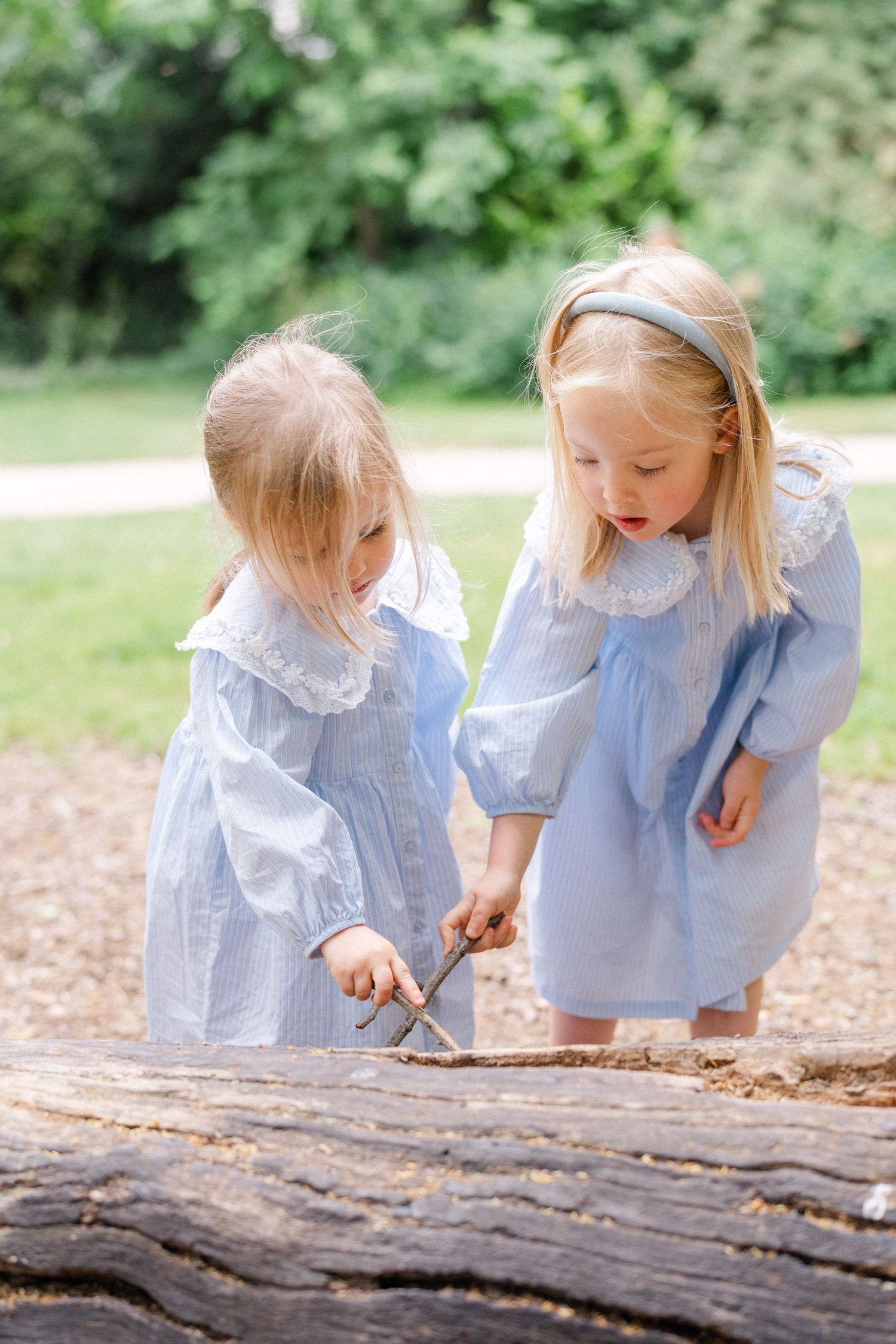Two young blonde girls in matching blue dresses playing with sticks on a log in a sunny park.