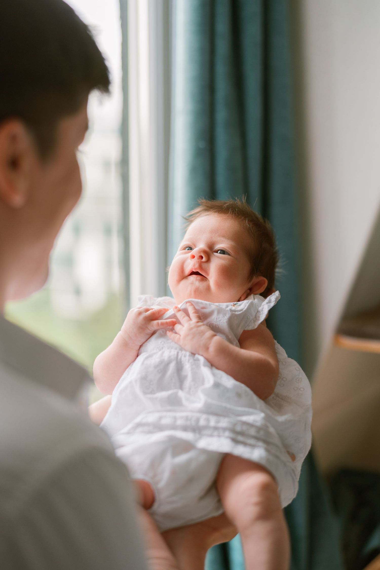 A smiling baby in a white dress being held up by a parent near a window with teal curtains.