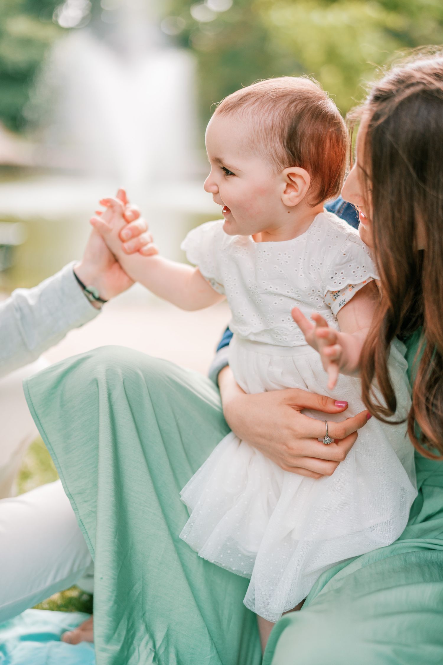 A joyful baby in a white dress sits on a mother's lap outdoors near a fountain on a sunny day.