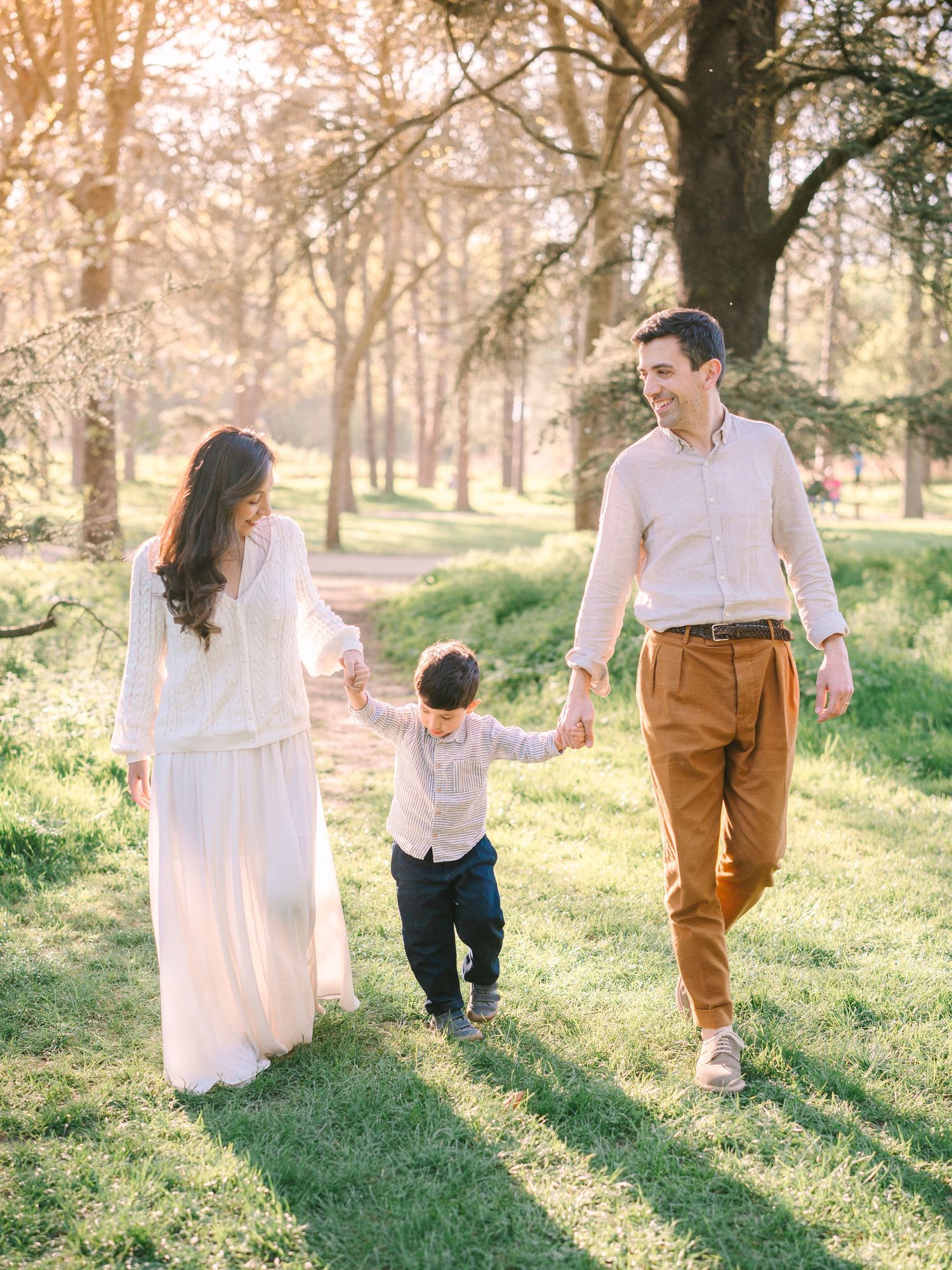 A young family walks hand-in-hand with their toddler through a sunlit park, wearing soft neutral and earth-toned outfits.