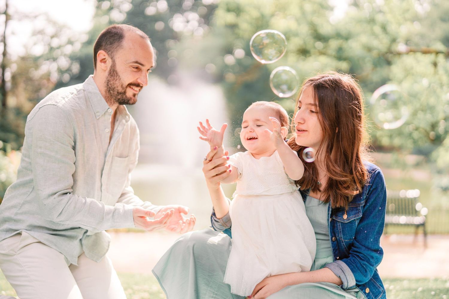 Happy family with baby girl playing with soap bubbles in sunny park, joyful parenting moment outdoors.