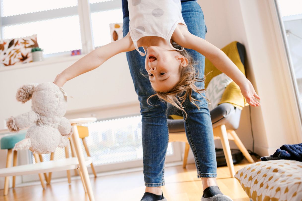 Adult playfully holding a laughing child upside down in a bright, modern living room with wooden floors.