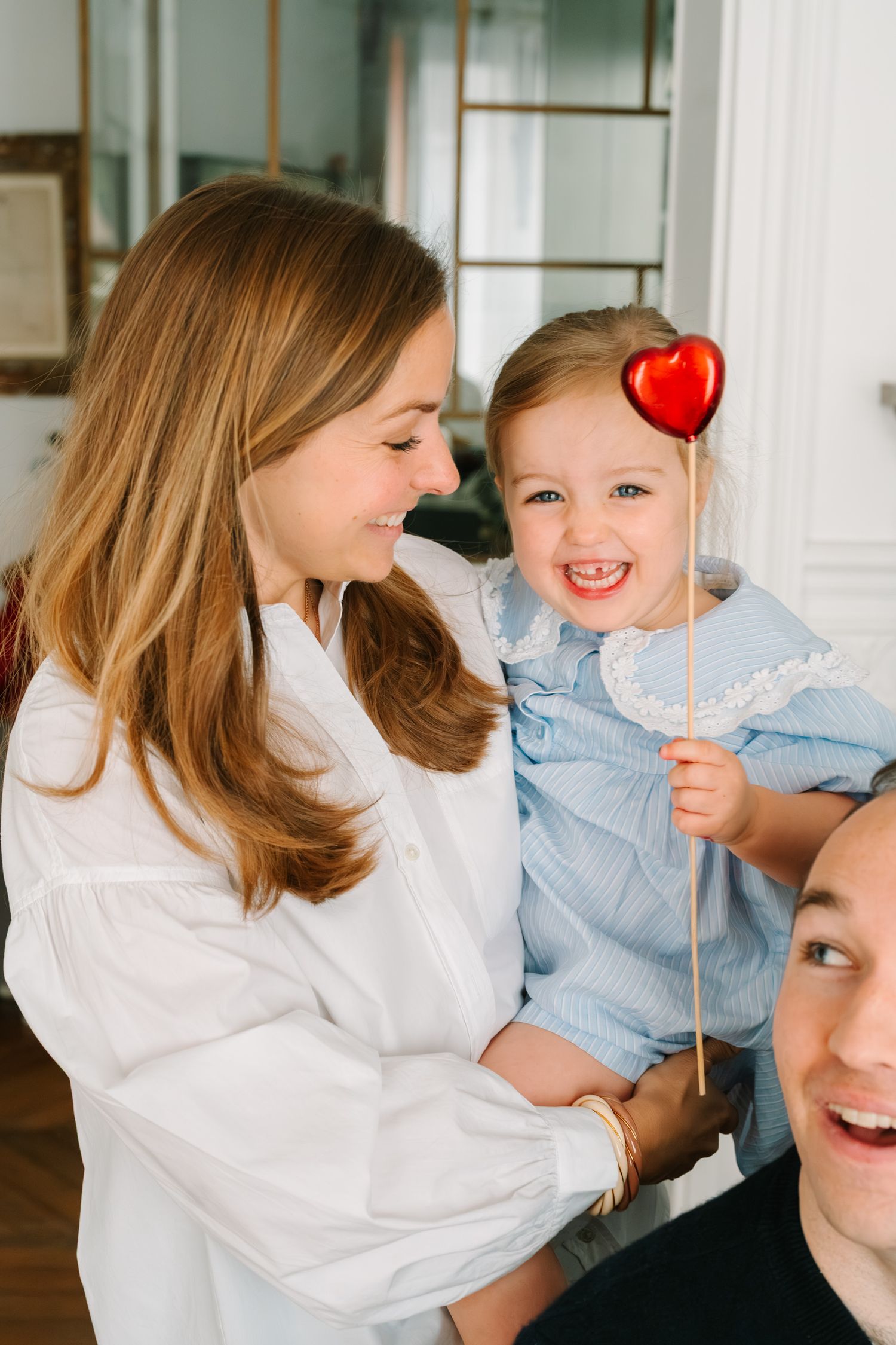 Mother in white top holds toddler girl in blue dress holding red heart lollipop.