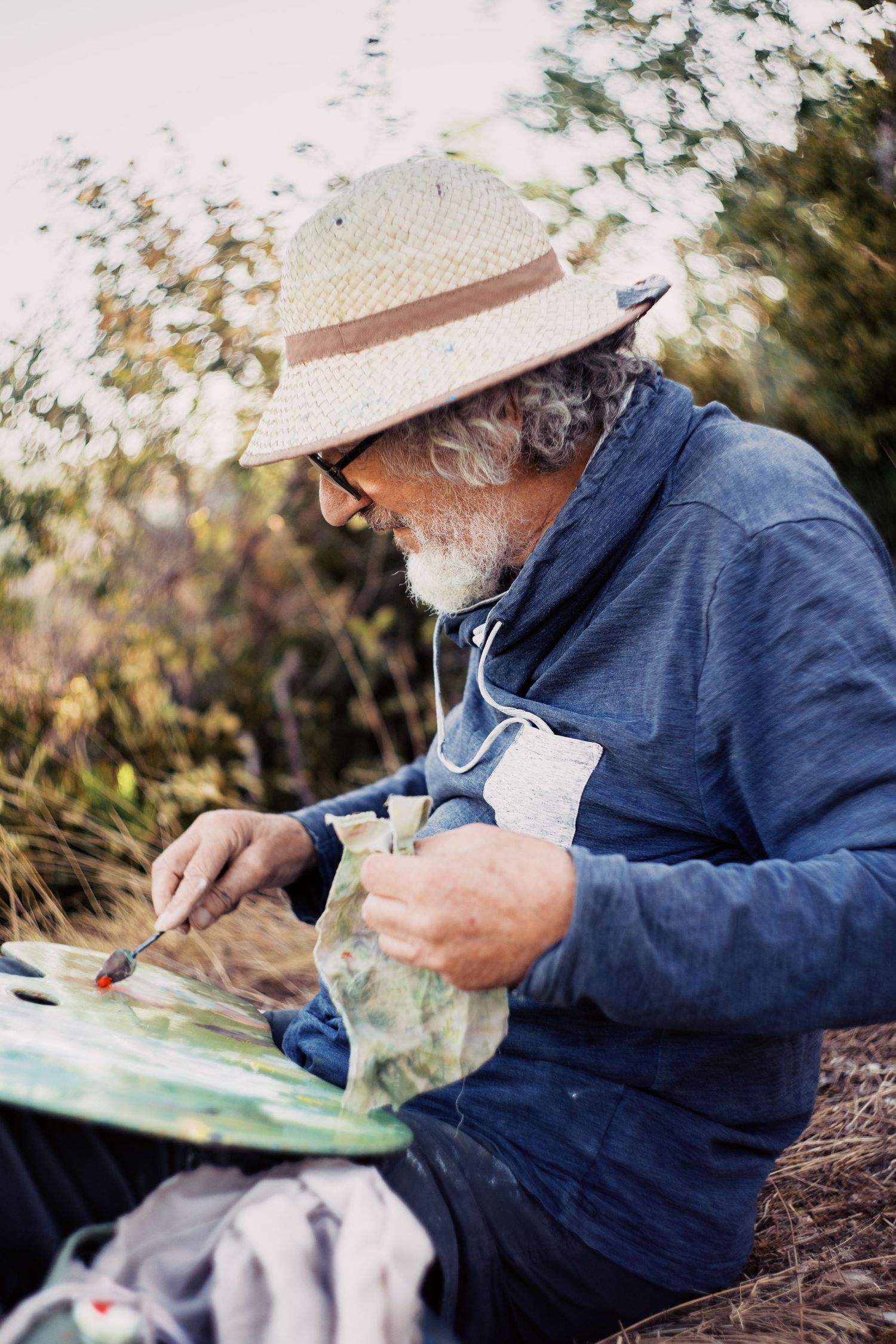 Elderly bearded artist in straw hat and glasses painting outdoors on canvas, surrounded by lush green foliage.