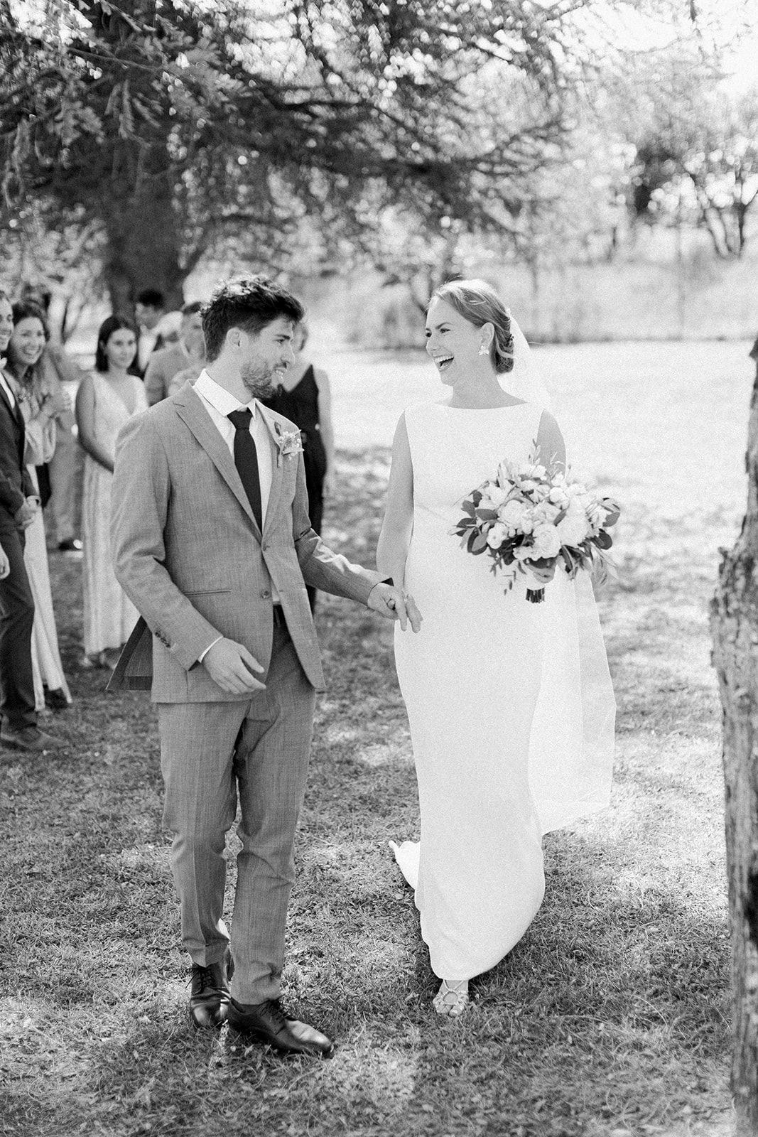 Black and white photo of a smiling bride holding bouquet walking with groom in gray suit at outdoor wedding ceremony.