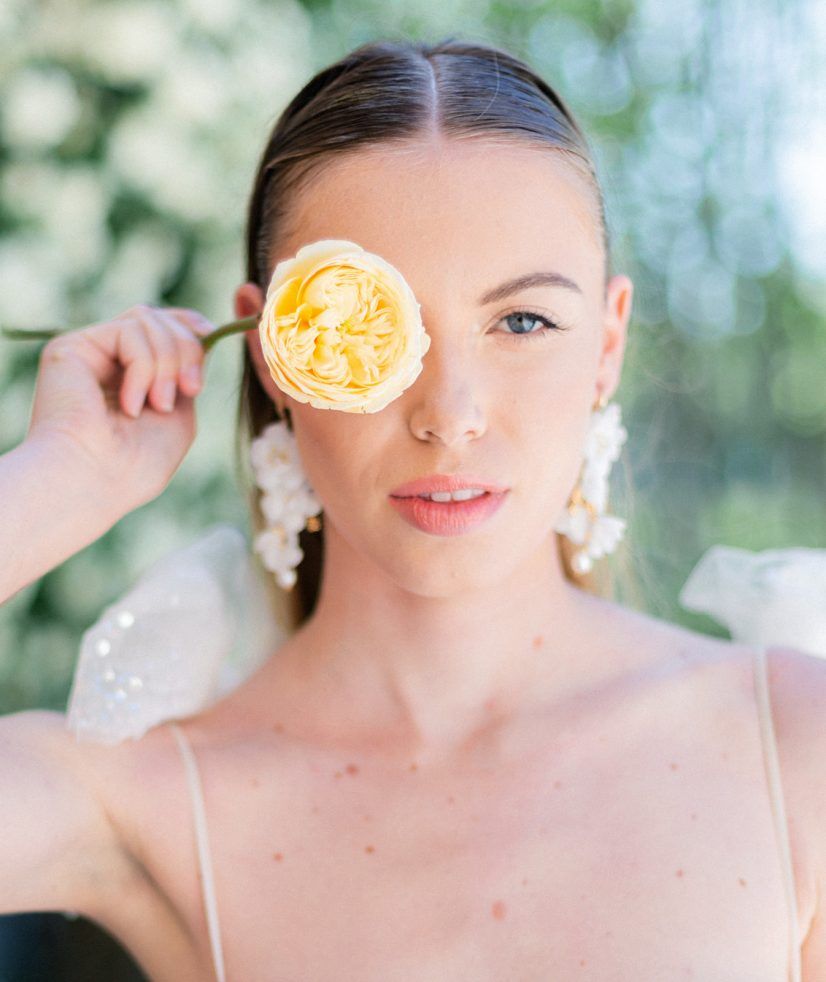 Elegant bride holding a yellow rose over one eye, wearing floral earrings and natural glam makeup.