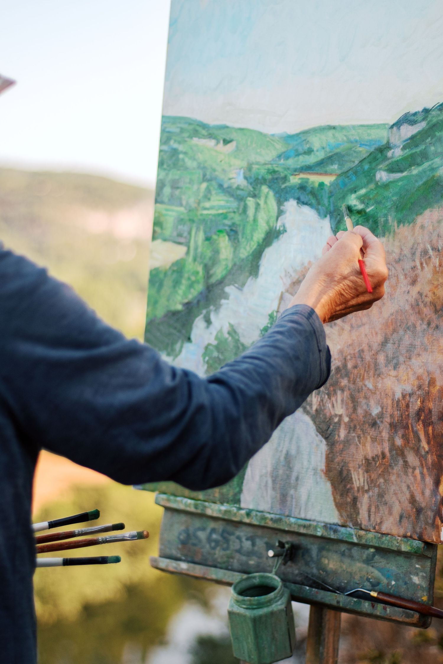 Artist painting a landscape on an easel outdoors, with brushes and green paint visible in natural light.