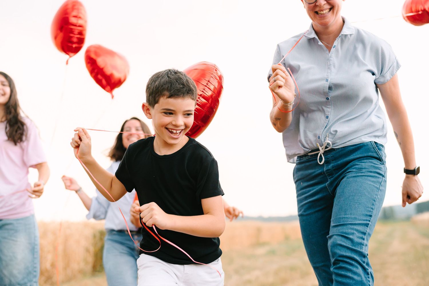 A joyful boy runs with red heart-shaped balloons alongside smiling women in a sunny field.