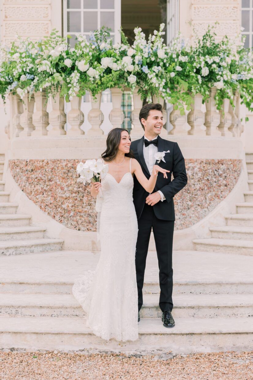 Bride and groom pose on elegant stone steps adorned with lush white floral arrangements at a classic wedding venue.
