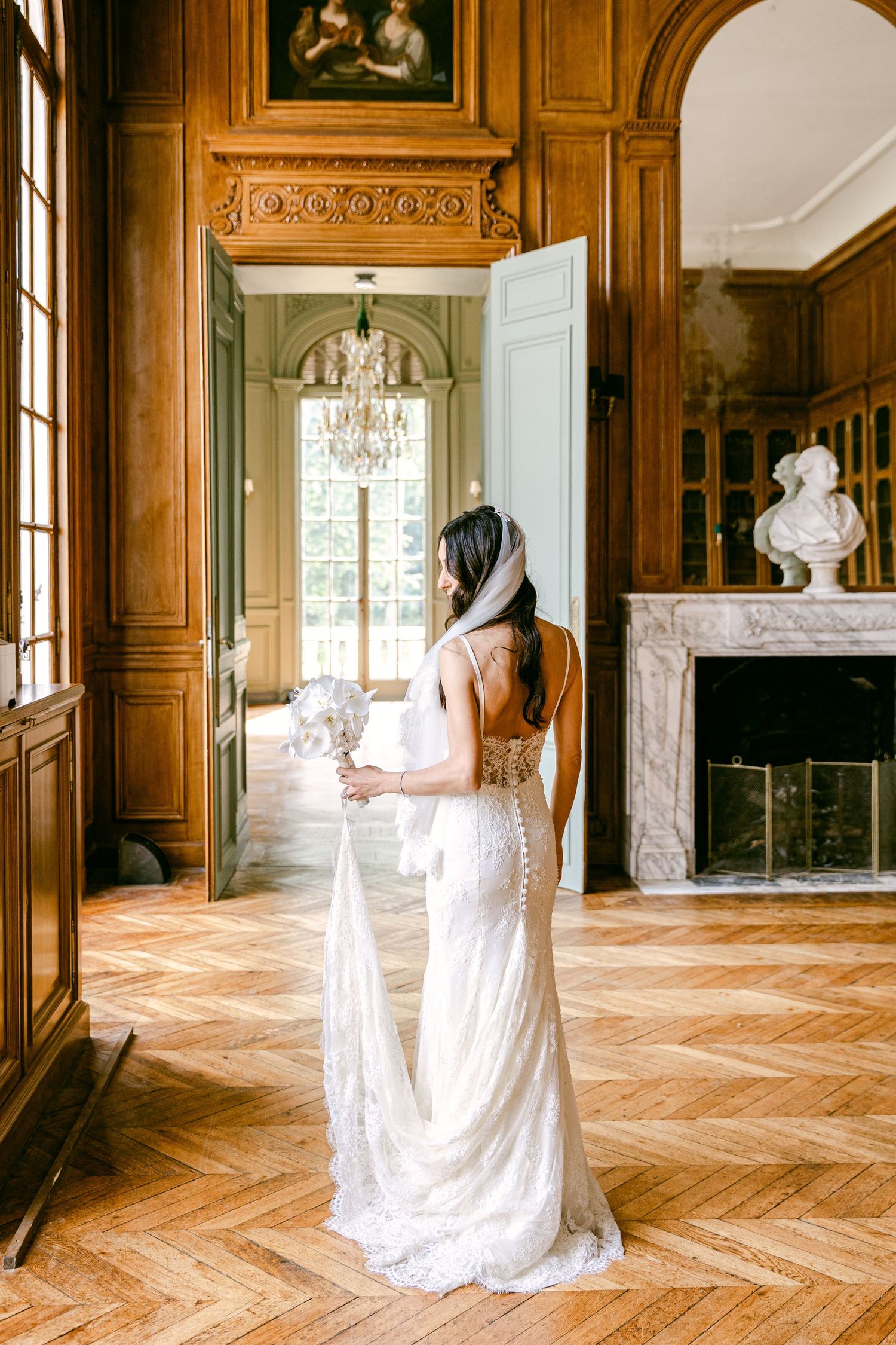 Bride in backless lace gown holding bouquet stands in elegant wood-paneled chateau with marble fireplace.