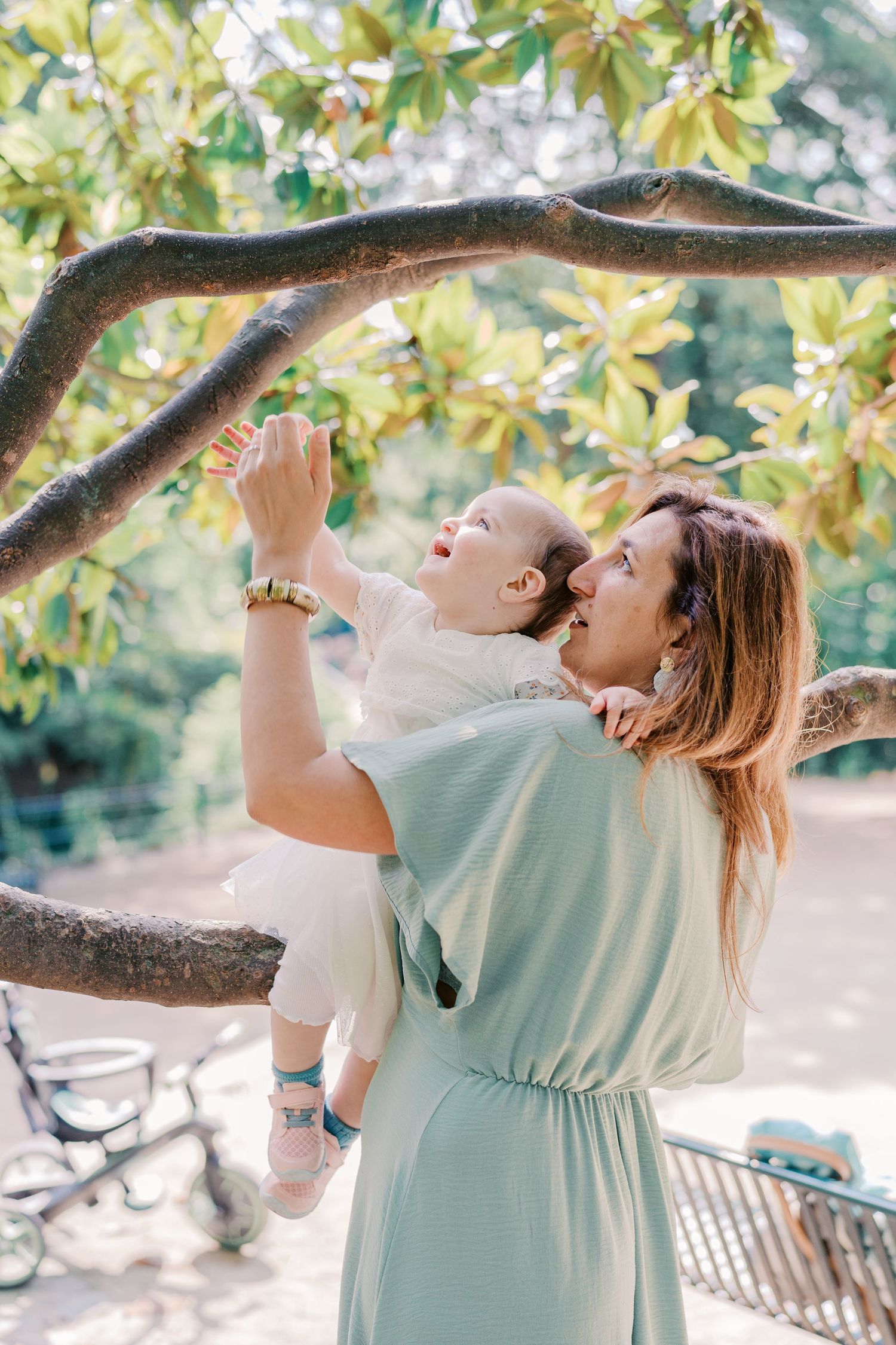 Parent soulevant son enfant pour attraper les branches d’un arbre dans un jardin baigné de lumière.
