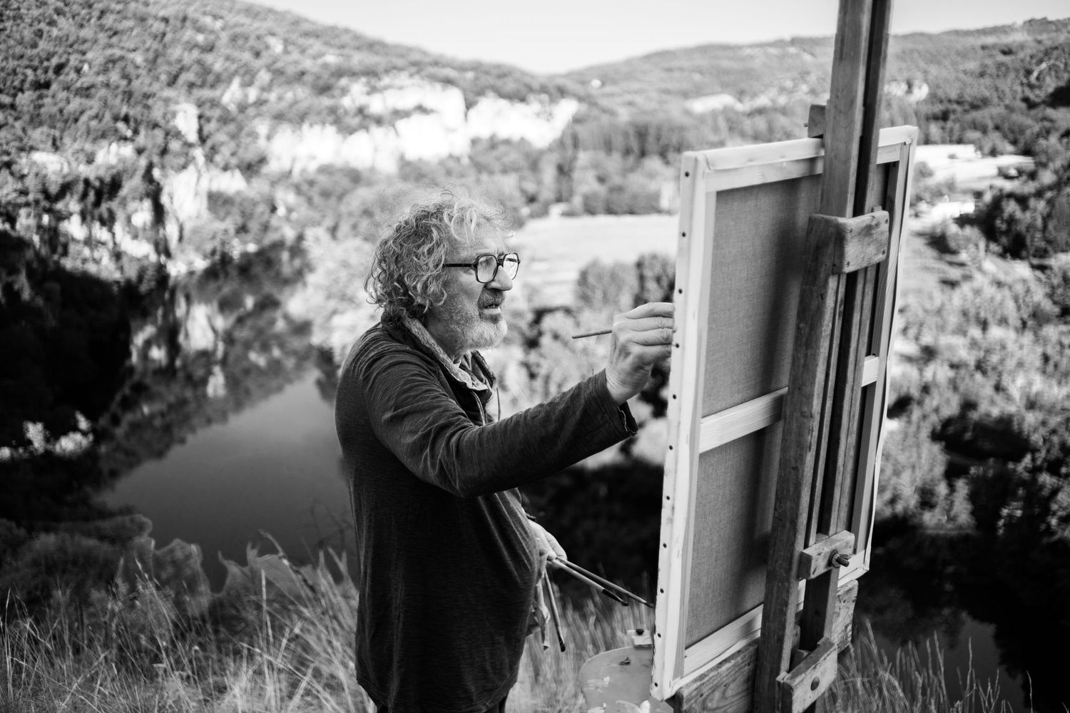 Black and white photo of an artist painting on canvas outdoors on a cliff overlooking a scenic river valley.