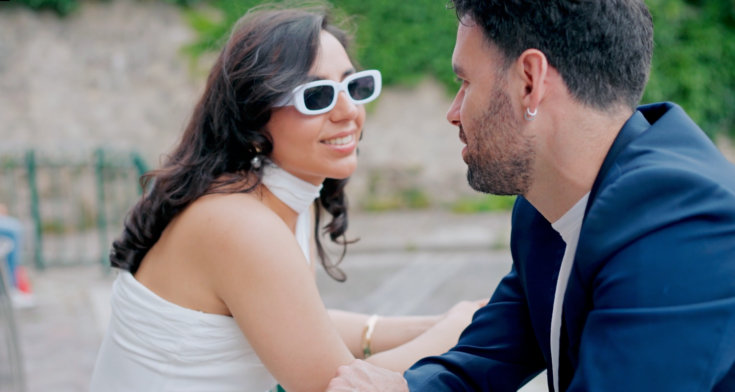 Couple sharing a romantic moment outdoors, woman in white dress and sunglasses smiling at man in navy suit.