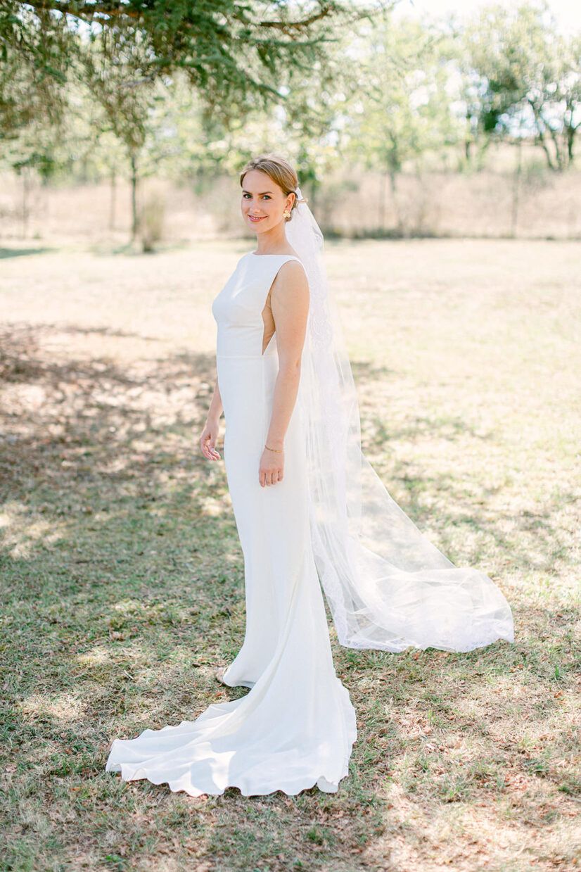 Bride in elegant white sleeveless gown with long veil stands outdoors in sunlit garden for bridal portrait.