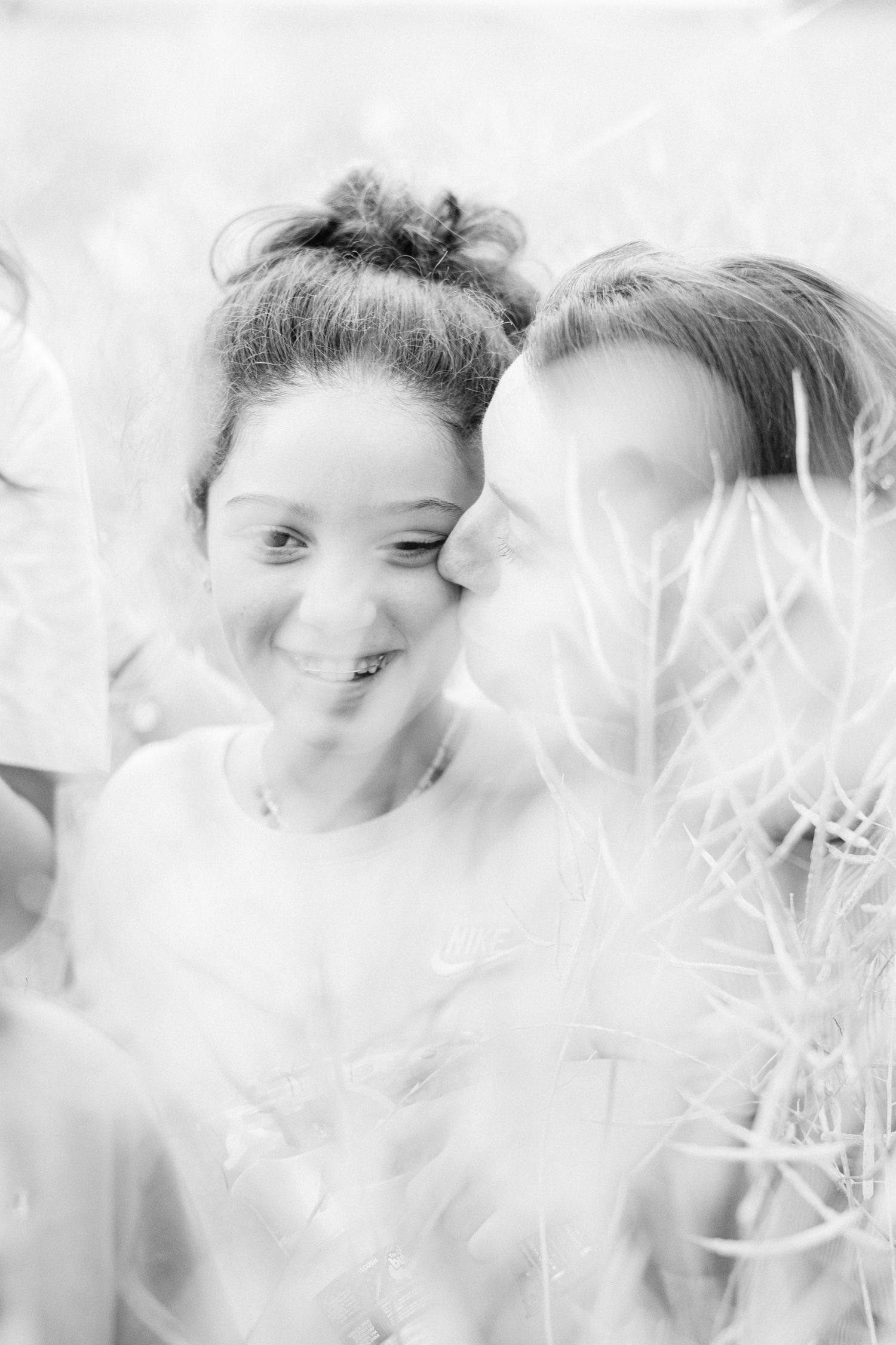 Black and white photo of two young women smiling outdoors, one kissing the other on the cheek among tall grass.