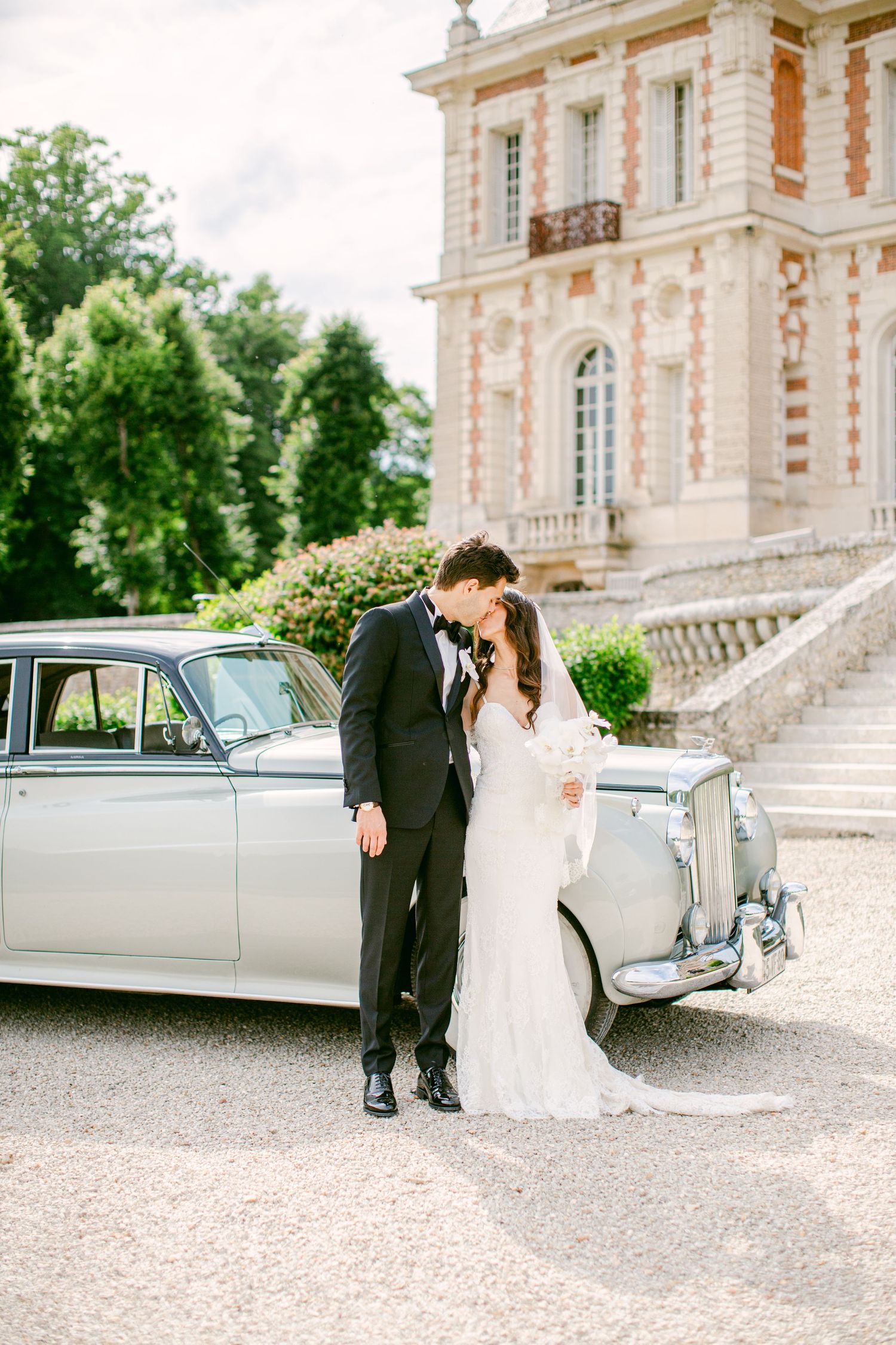 Bride and groom kissing beside a vintage white car in front of an elegant French chateau on their wedding day.