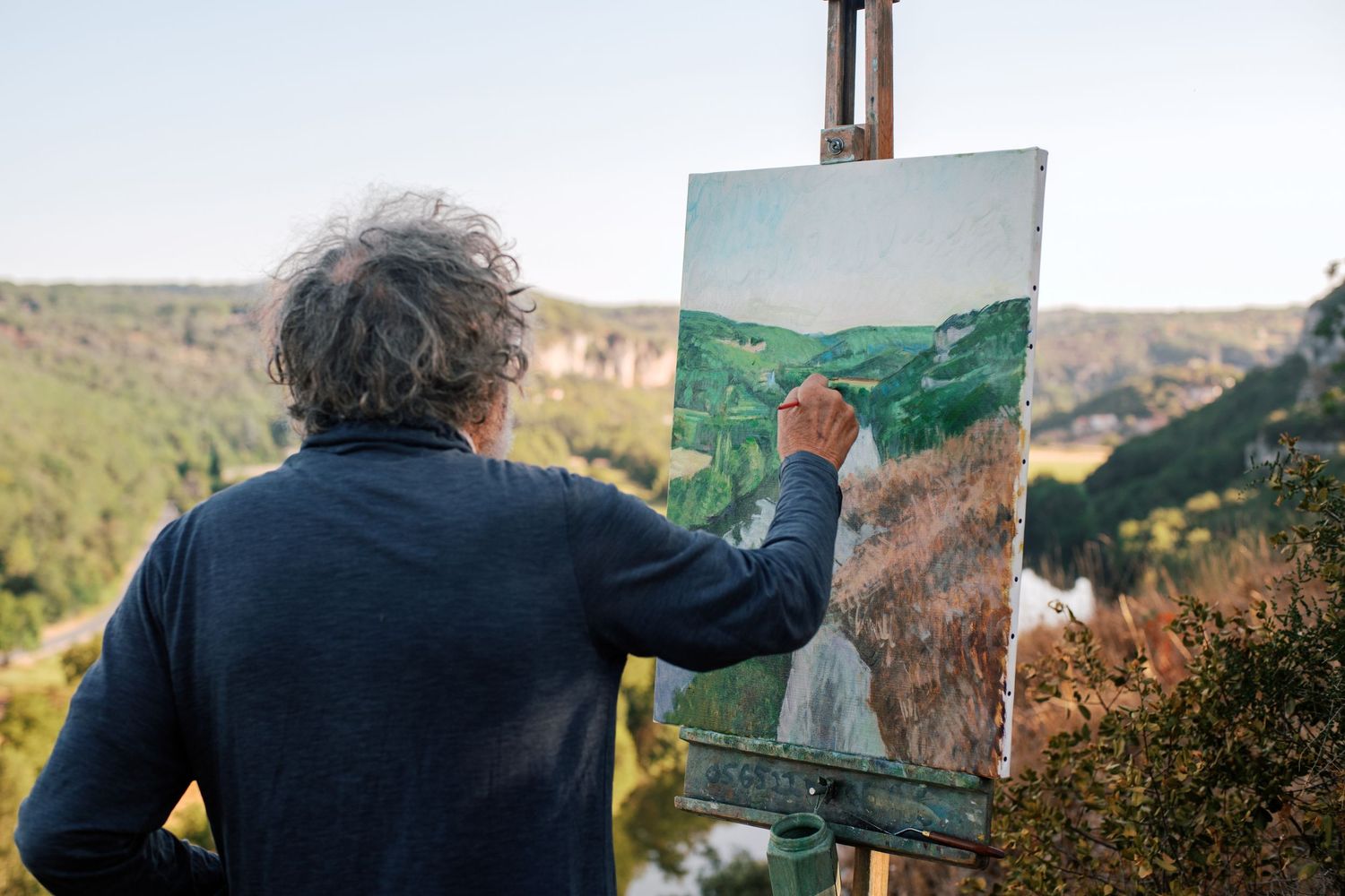 Artist painting a landscape on canvas outdoors on an easel with a scenic river valley view.