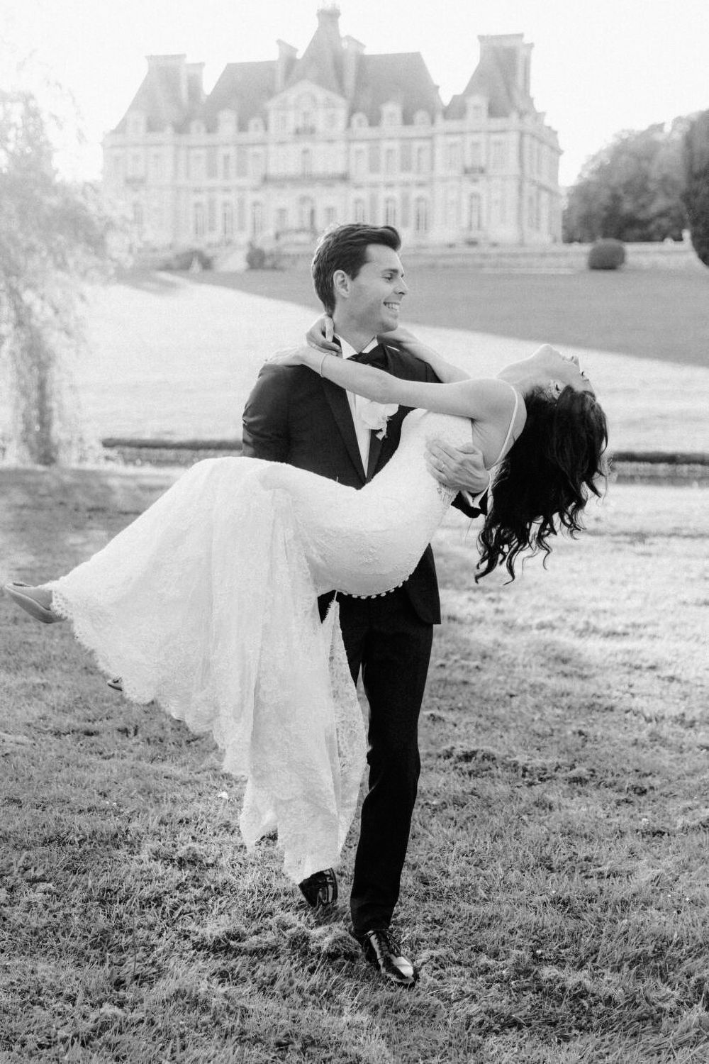Groom lifts bride in wedding dress outside a French chateau in black and white photo.