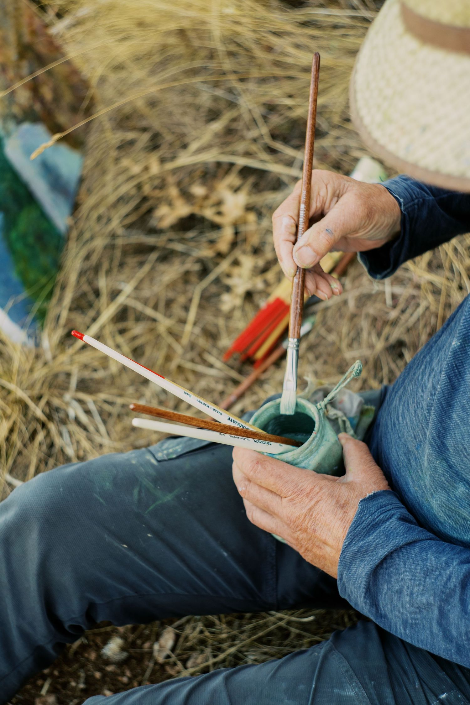 Artist sitting outdoors holding paintbrushes and a green cup, painting in a natural field setting while wearing a straw hat.