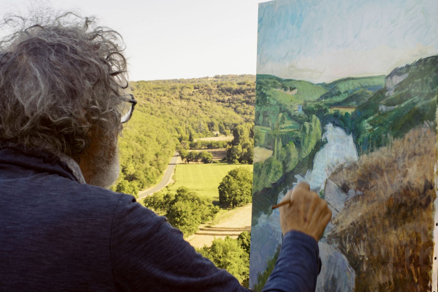 Artist painting a lush green valley landscape on canvas outdoors, capturing rolling hills and a winding river below.