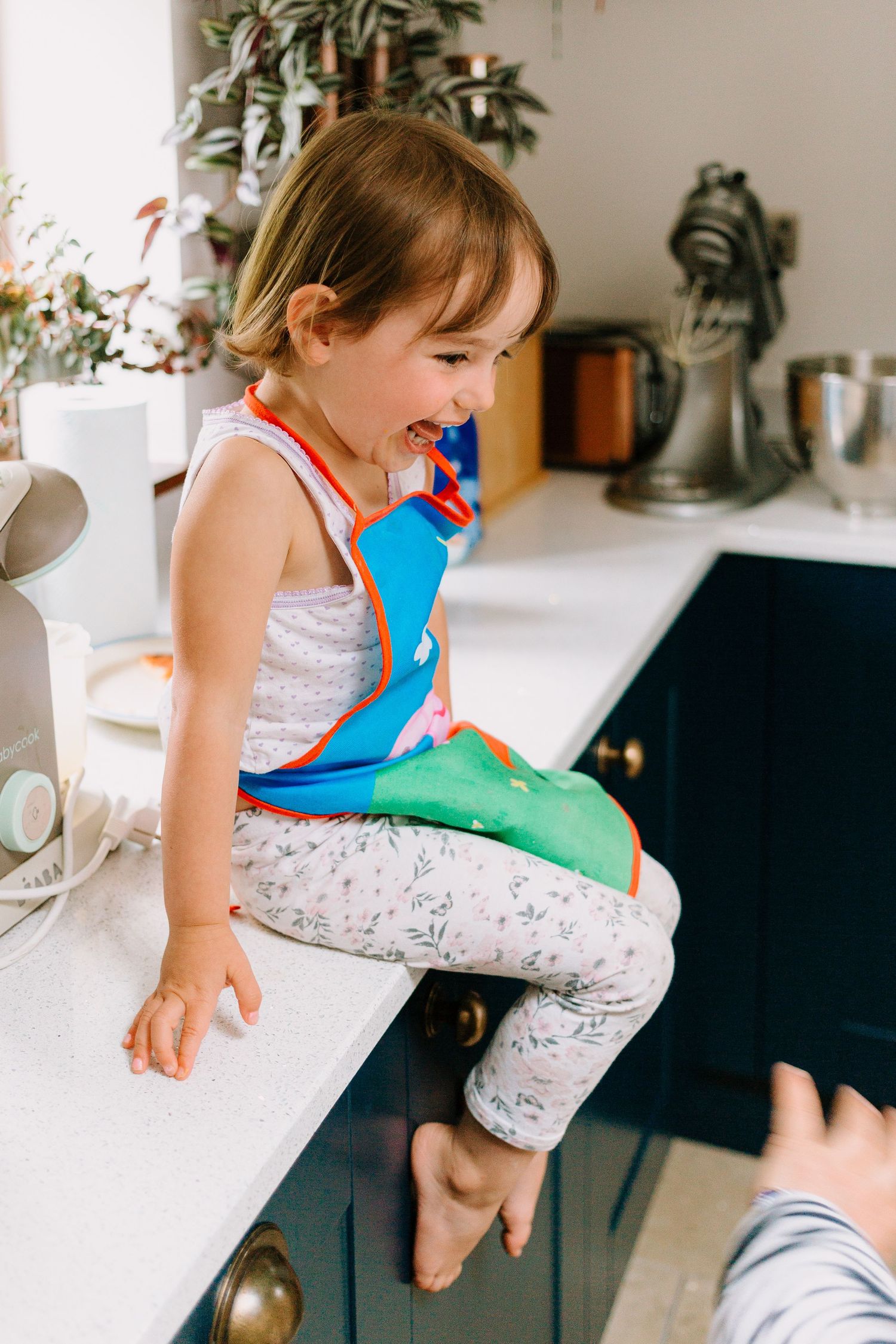 Happy toddler girl sitting on kitchen counter wearing colorful apron, laughing in bright modern kitchen.