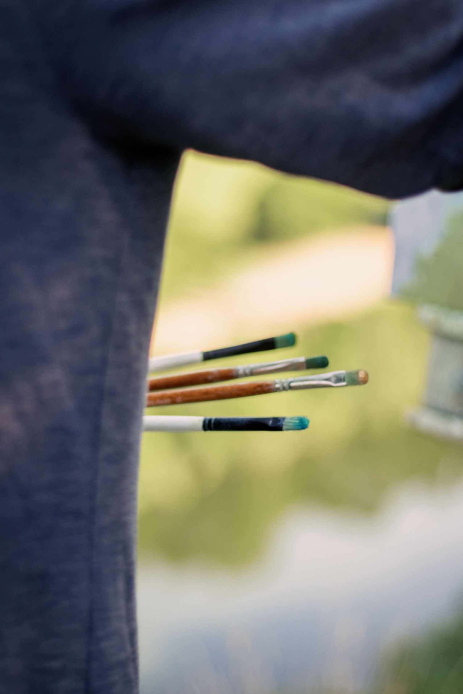 Artist holding multiple paint brushes with green paint outdoors, blurred natural background.