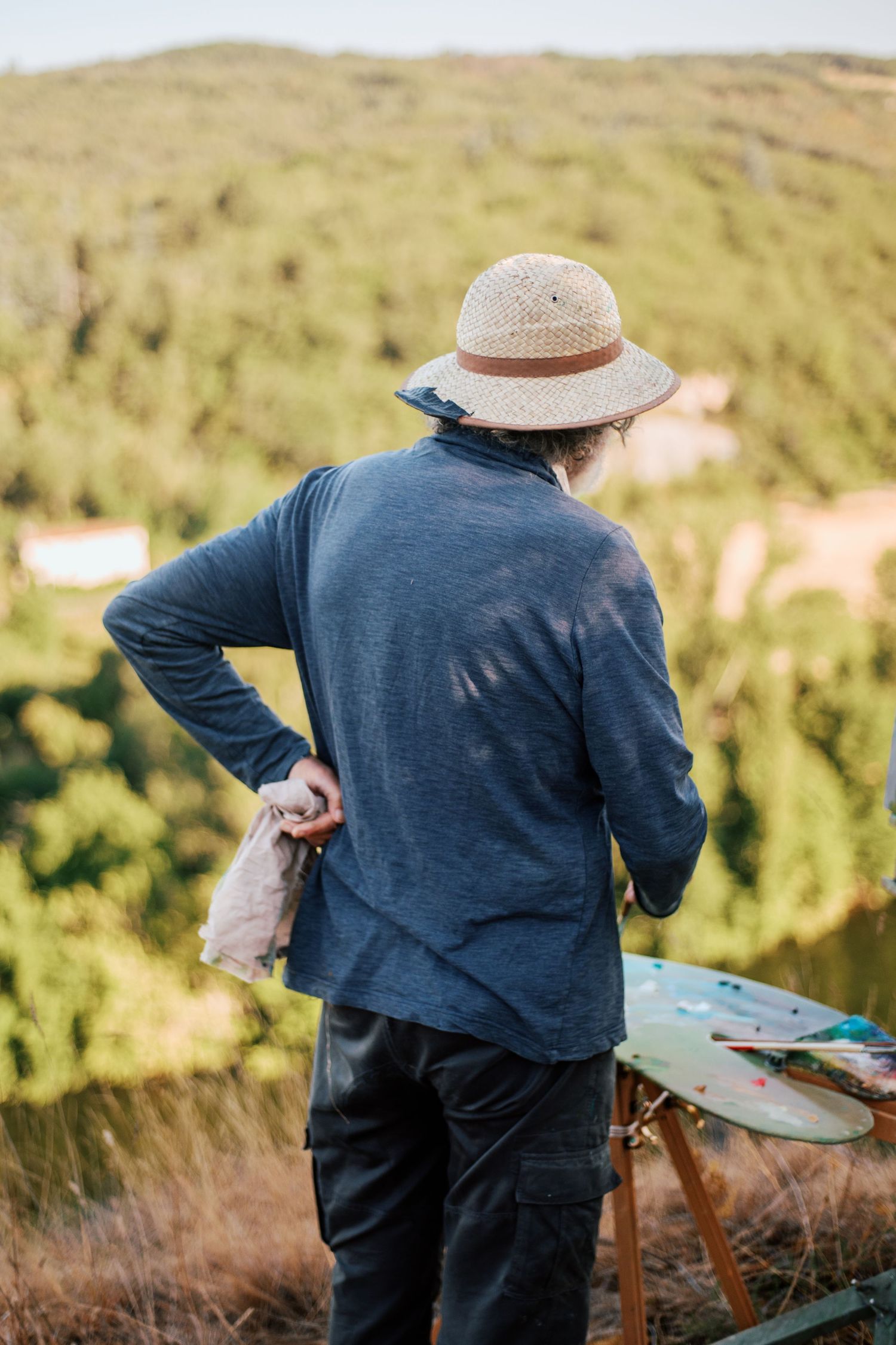 Artist in straw hat painting outdoors on an easel, surrounded by lush green hills and natural landscape.