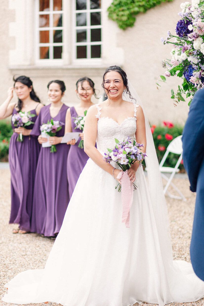Smiling bride in white gown holds purple bouquet, bridesmaids in purple dresses stand behind at outdoor ceremony.