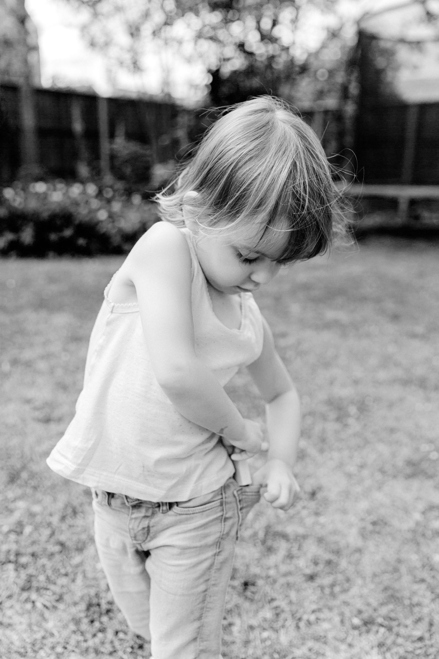 Black and white photo of a young toddler girl in a tank top and jeans playing outdoors in a backyard.