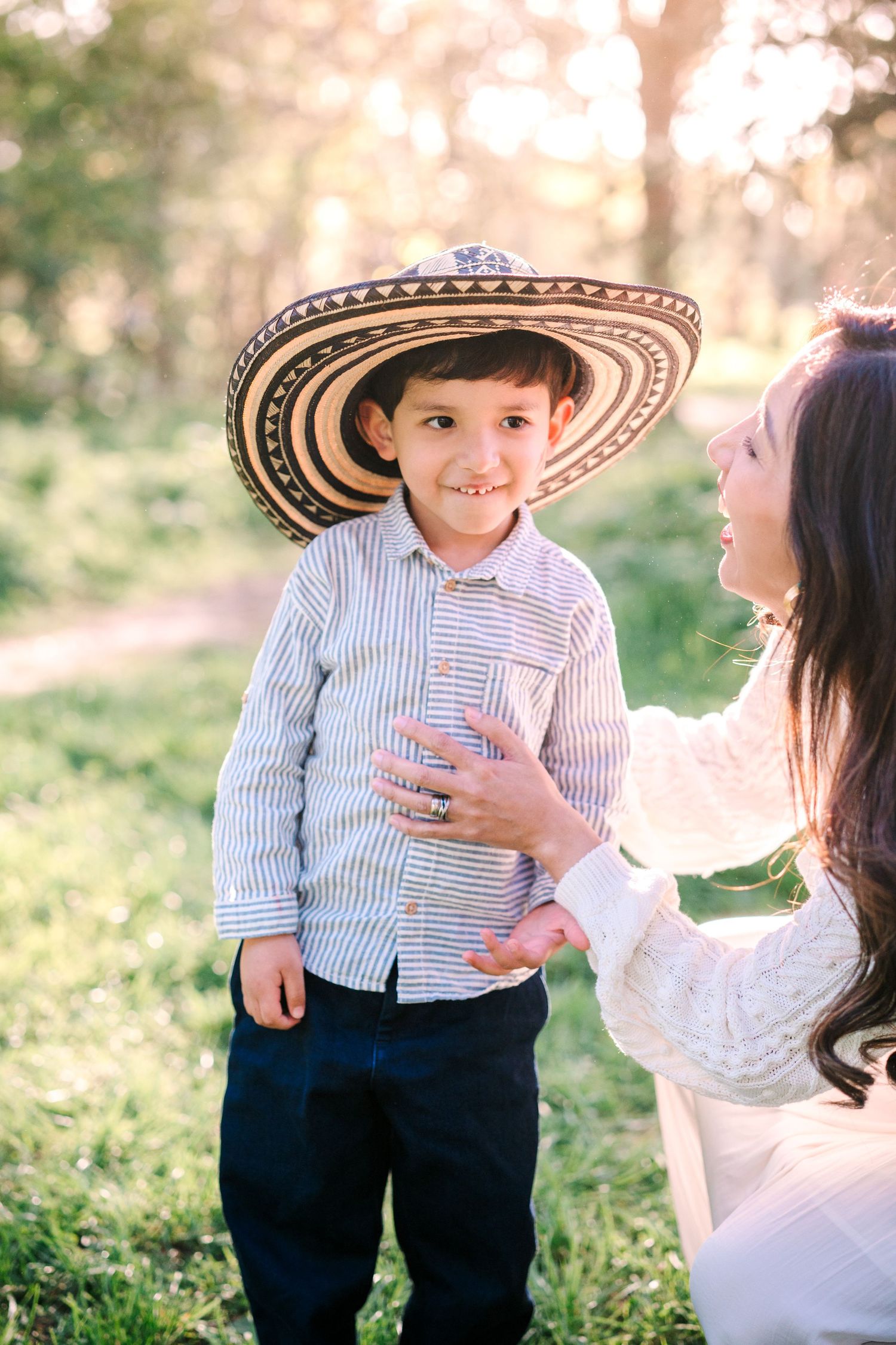 A young boy wearing a sombrero and striped shirt smiles outdoors while a woman in white dress adjusts his clothing.
