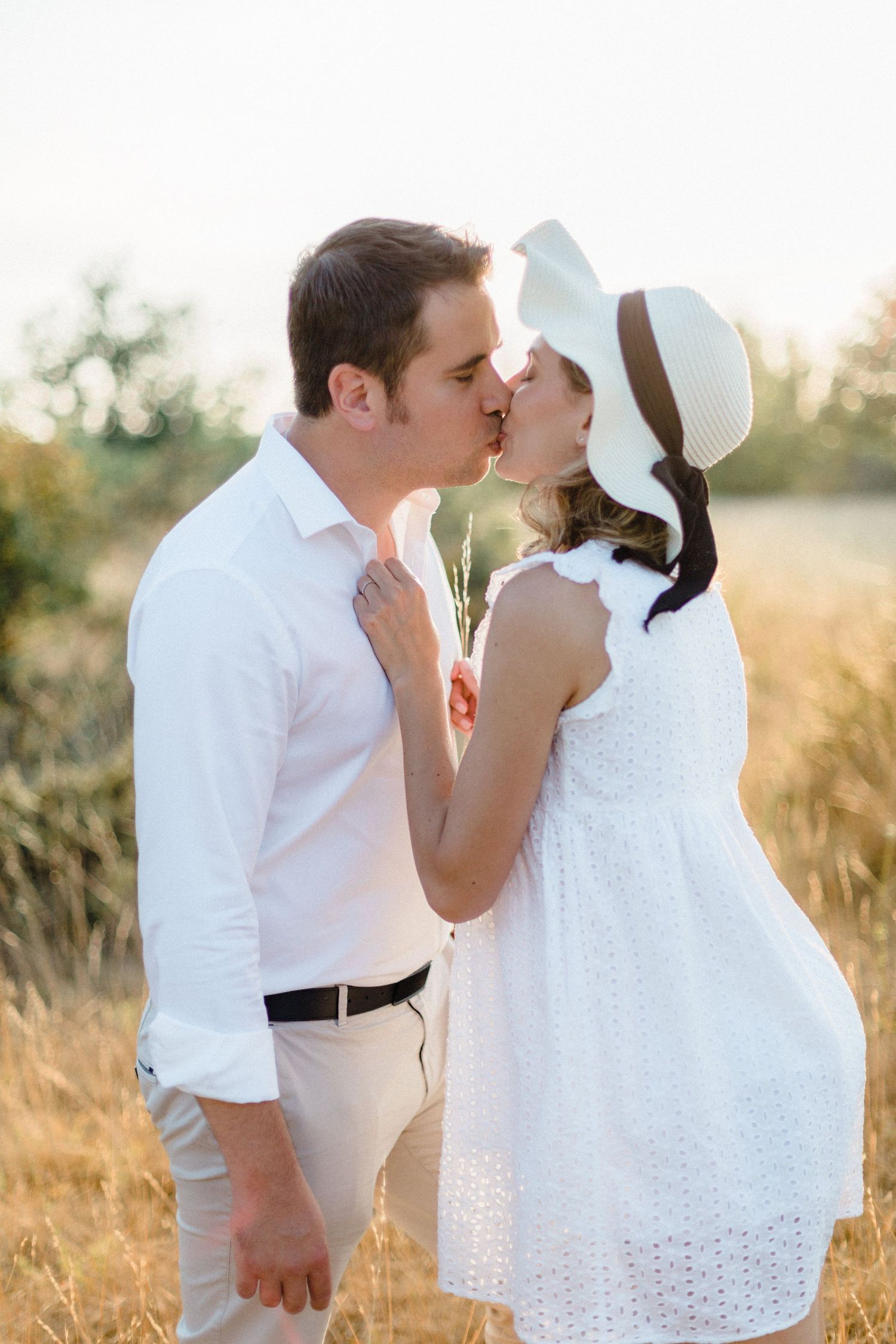 Romantic outdoor moment at sunset featuring couple in white attire sharing a tender kiss in a golden field.