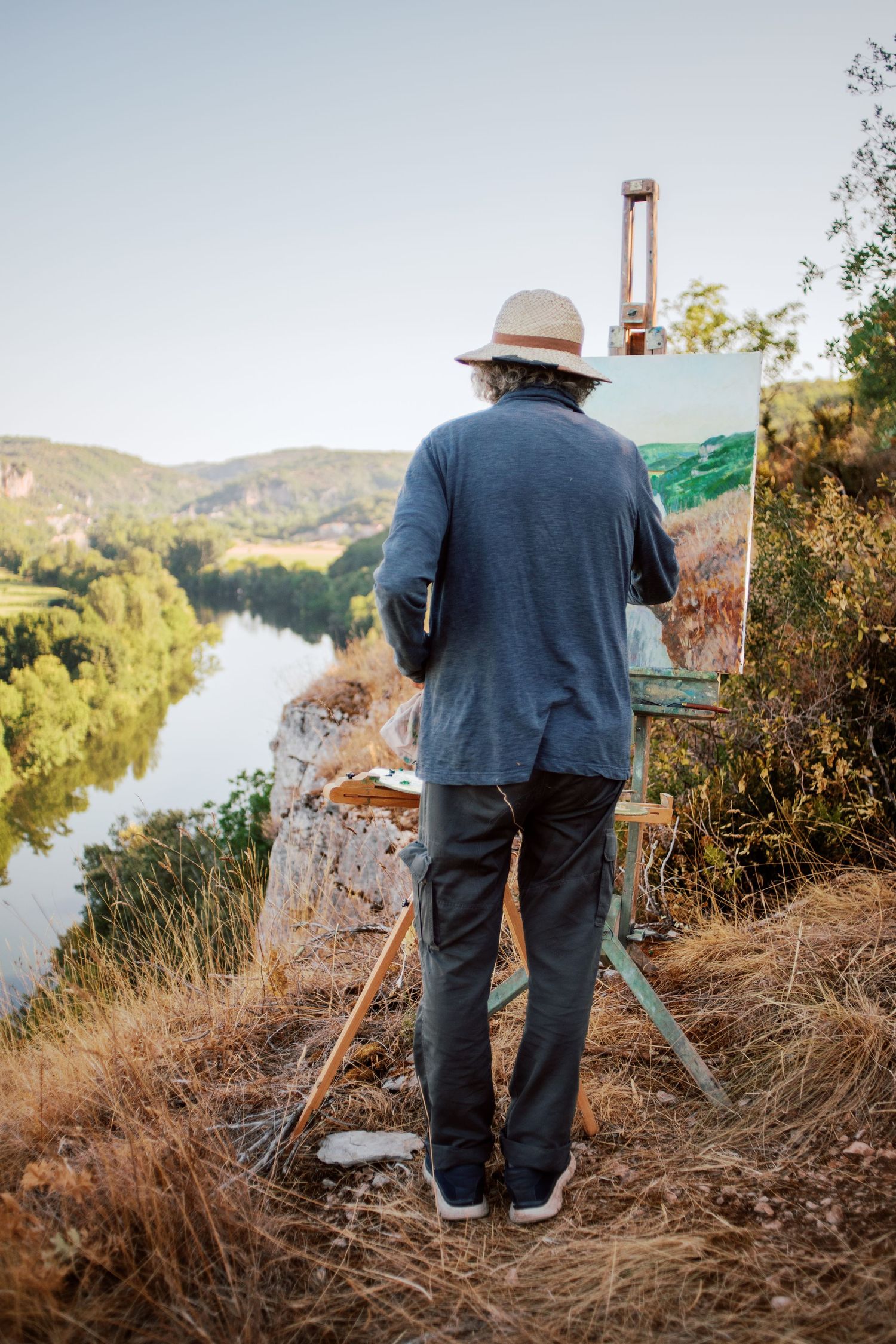 Artist painting landscape on easel outdoors, overlooking a scenic river valley with lush green hills under a clear sky.