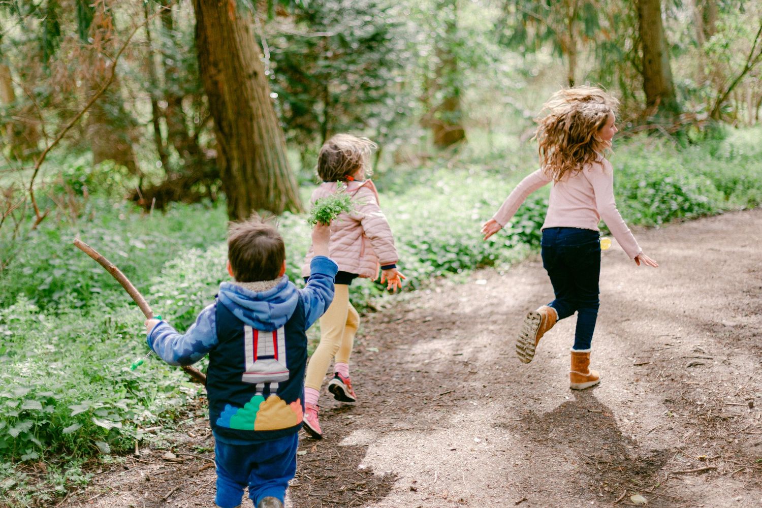 Children in colorful clothing walk along a winding dirt trail through a lush green forest on a summer day.