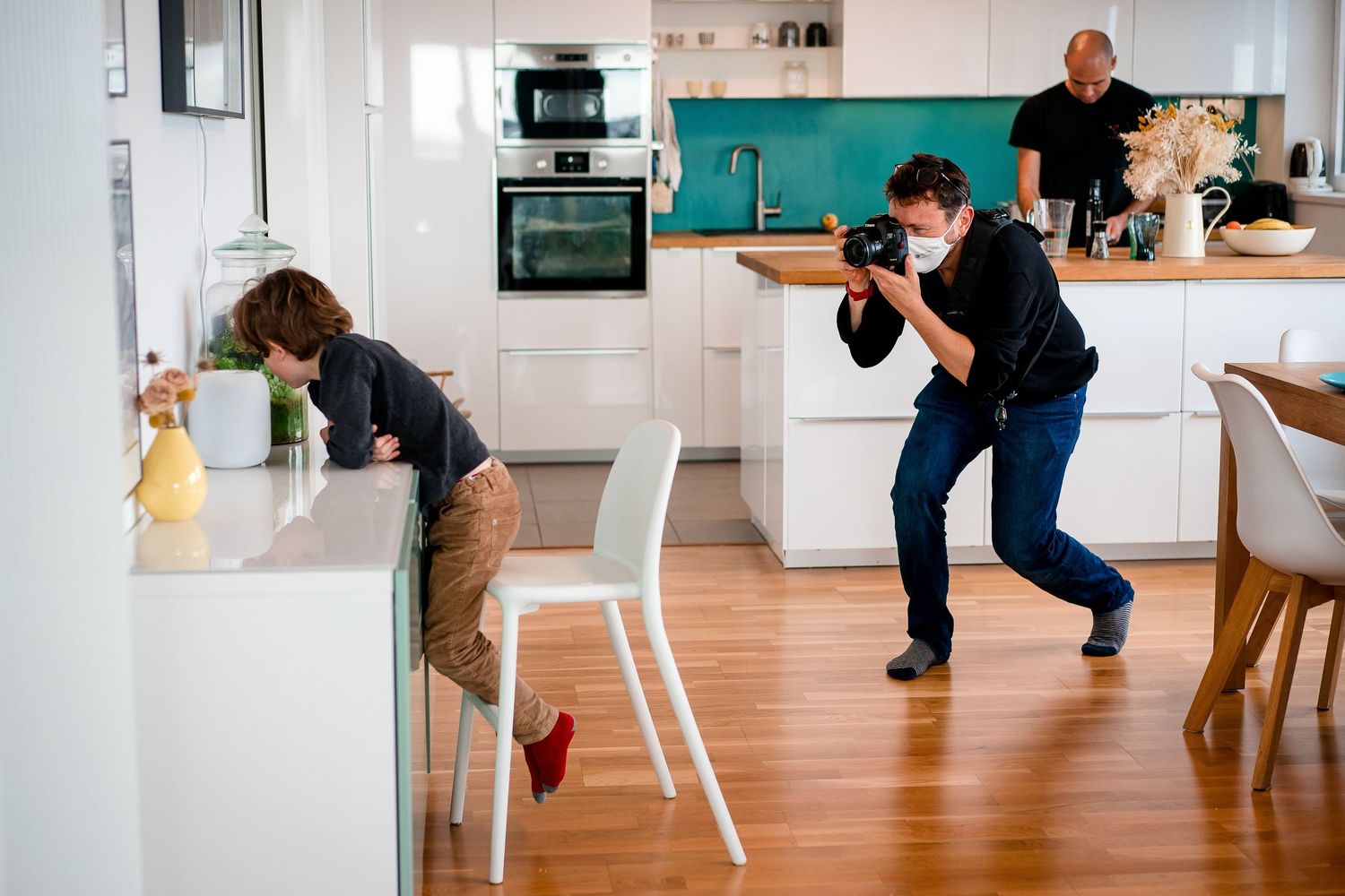Photographer crouching to capture a candid shot of a child in a modern white kitchen with wooden floors.