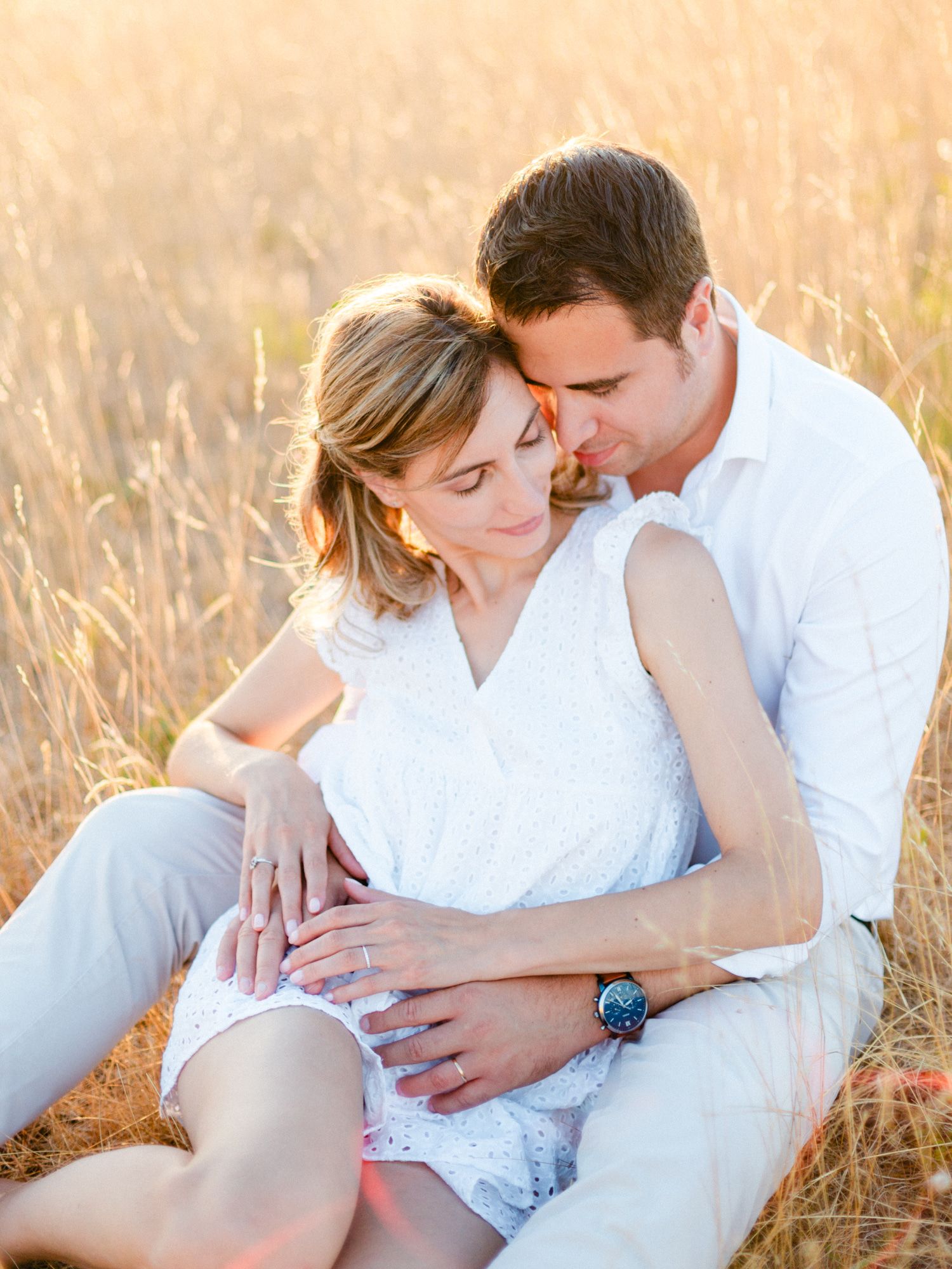 Couple sitting in golden wheat field at sunset, woman in white dress embraced by man in white shirt.