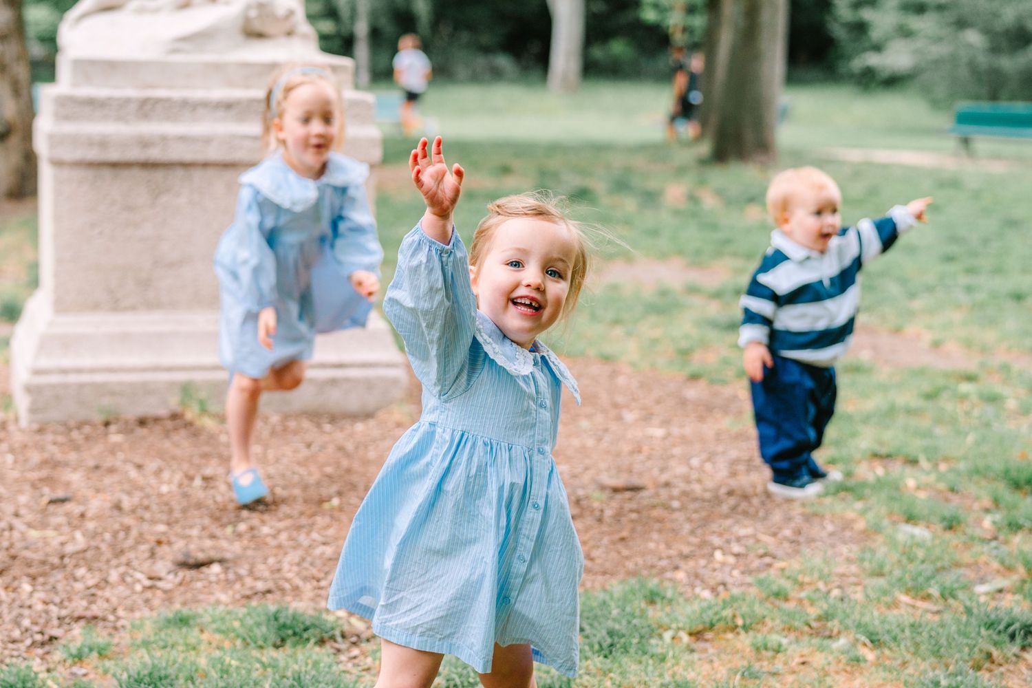 Three young children playing outdoors in a park, wearing blue outfits, with a girl in front laughing and reaching up.