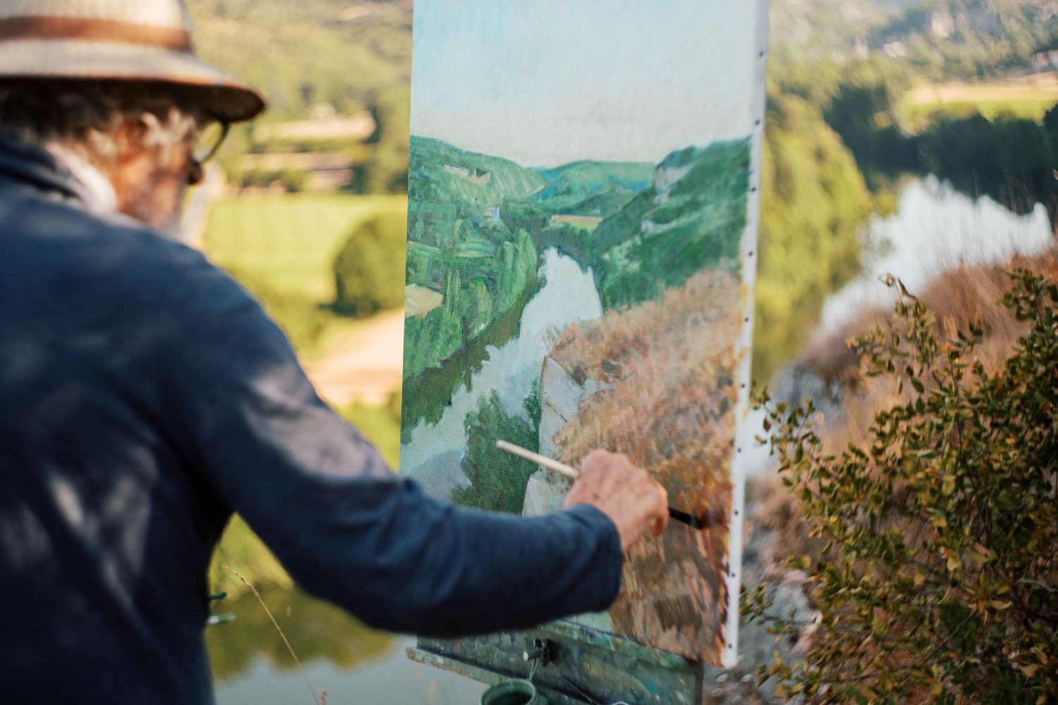 Artist painting a lush river valley landscape on canvas at an outdoor easel in natural sunlight.