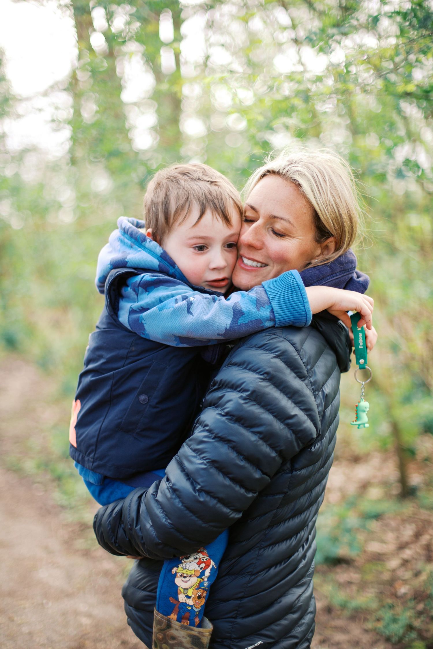A smiling woman in a blue puffer jacket hugs a young boy in a forest setting.